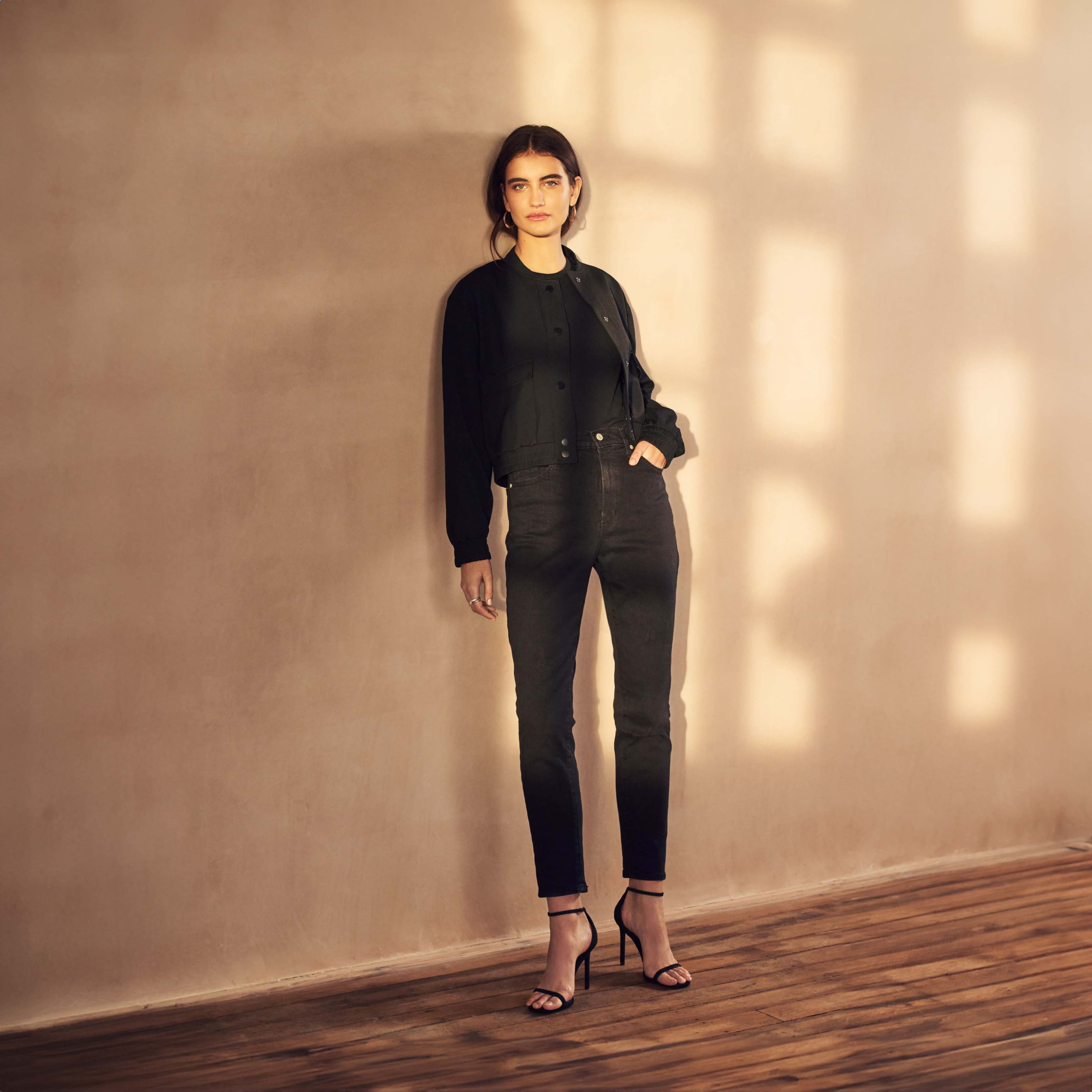 A person stands against a softly lit, neutral-toned wall, wearing a black cropped jacket and slim black pants, with strappy black heels on a wooden floor.