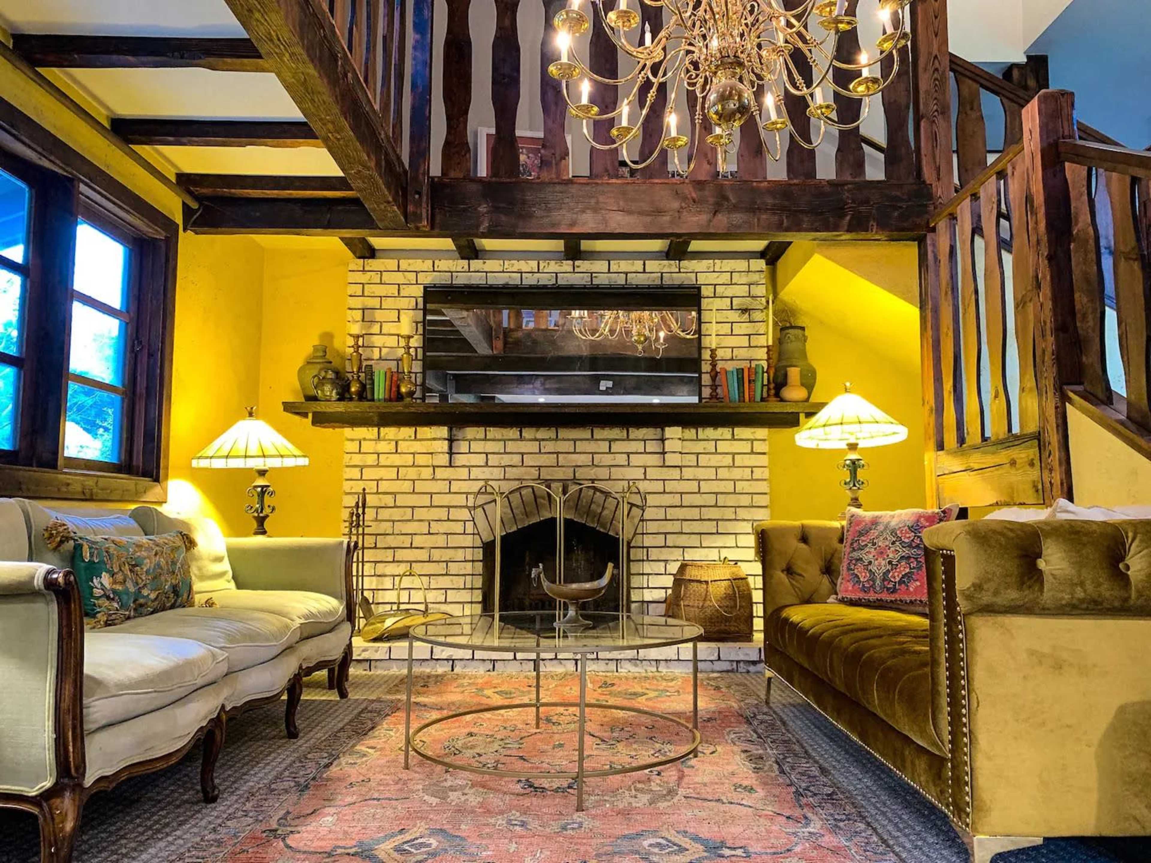 The image depicts a cozy living room with a brick fireplace, a chandelier, two sofas, and a patterned rug, set against yellow walls.
