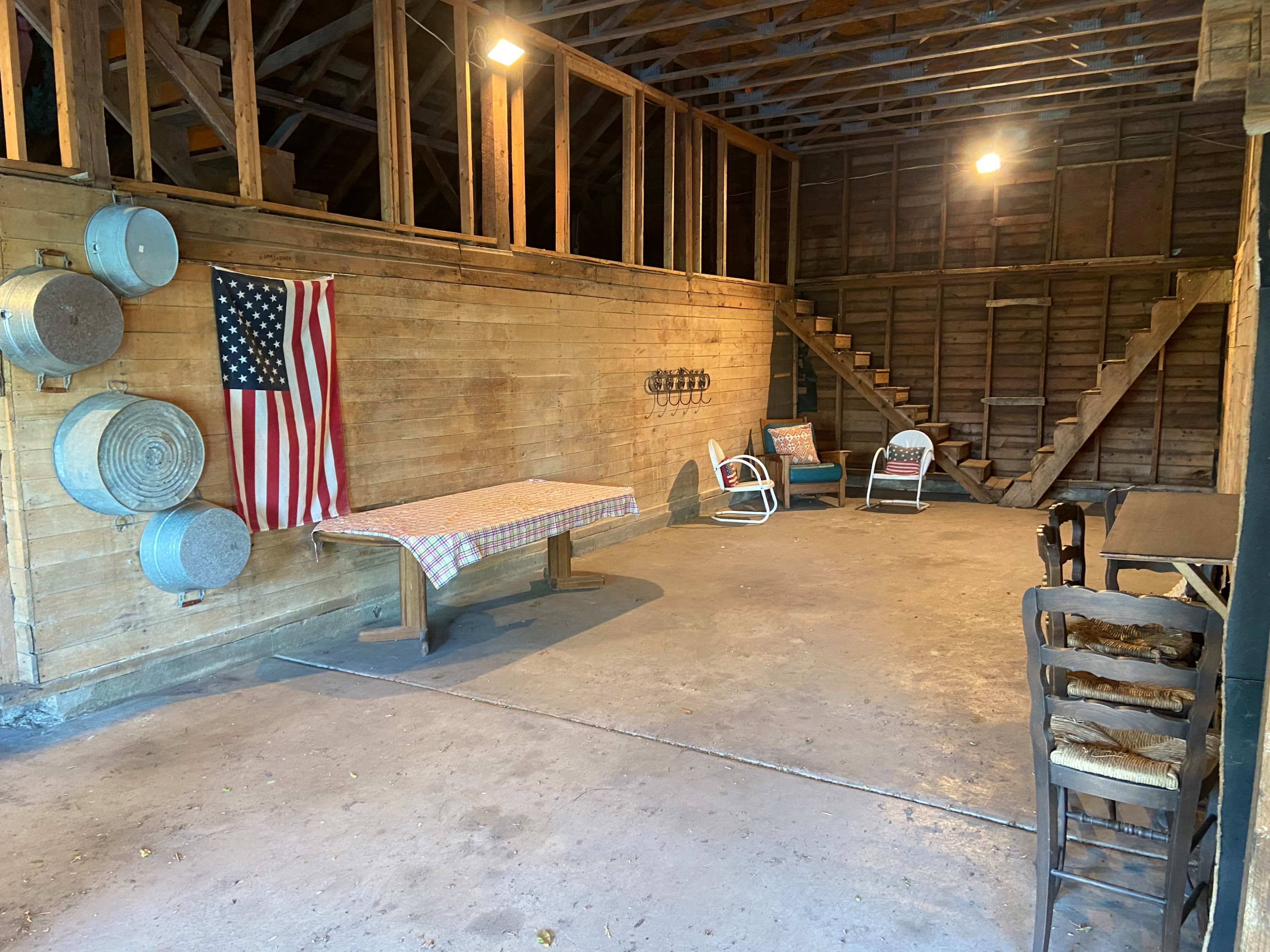 An interior of a wooden barn features a table with a checkered tablecloth, two chairs, an American flag hanging on the wall, and a staircase leading to an upper level.