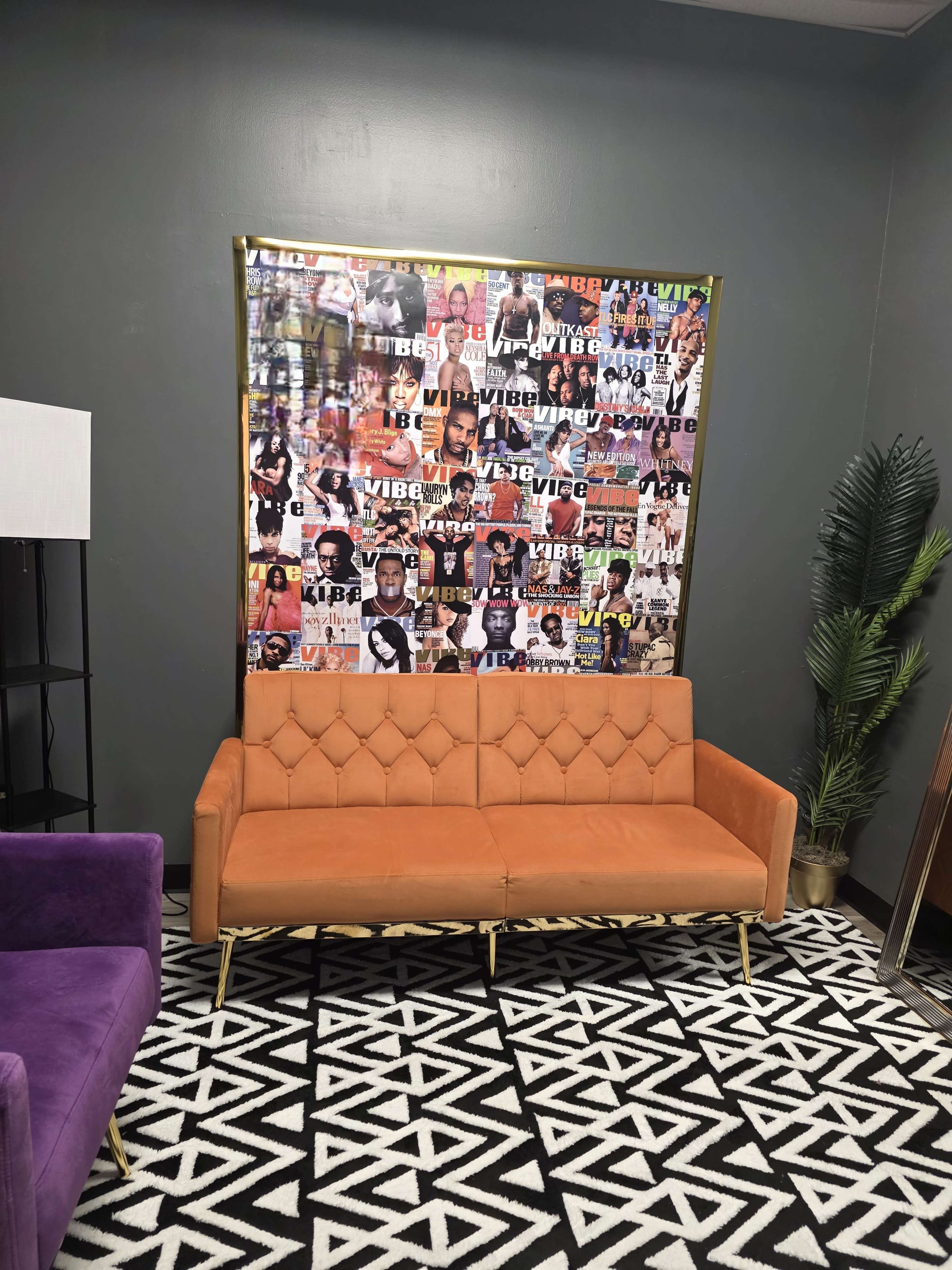 The image shows a modern lounge area featuring an orange tufted sofa, a patterned black and white rug, a purple chair, a standing lamp, and a large collage of magazine covers on the wall.