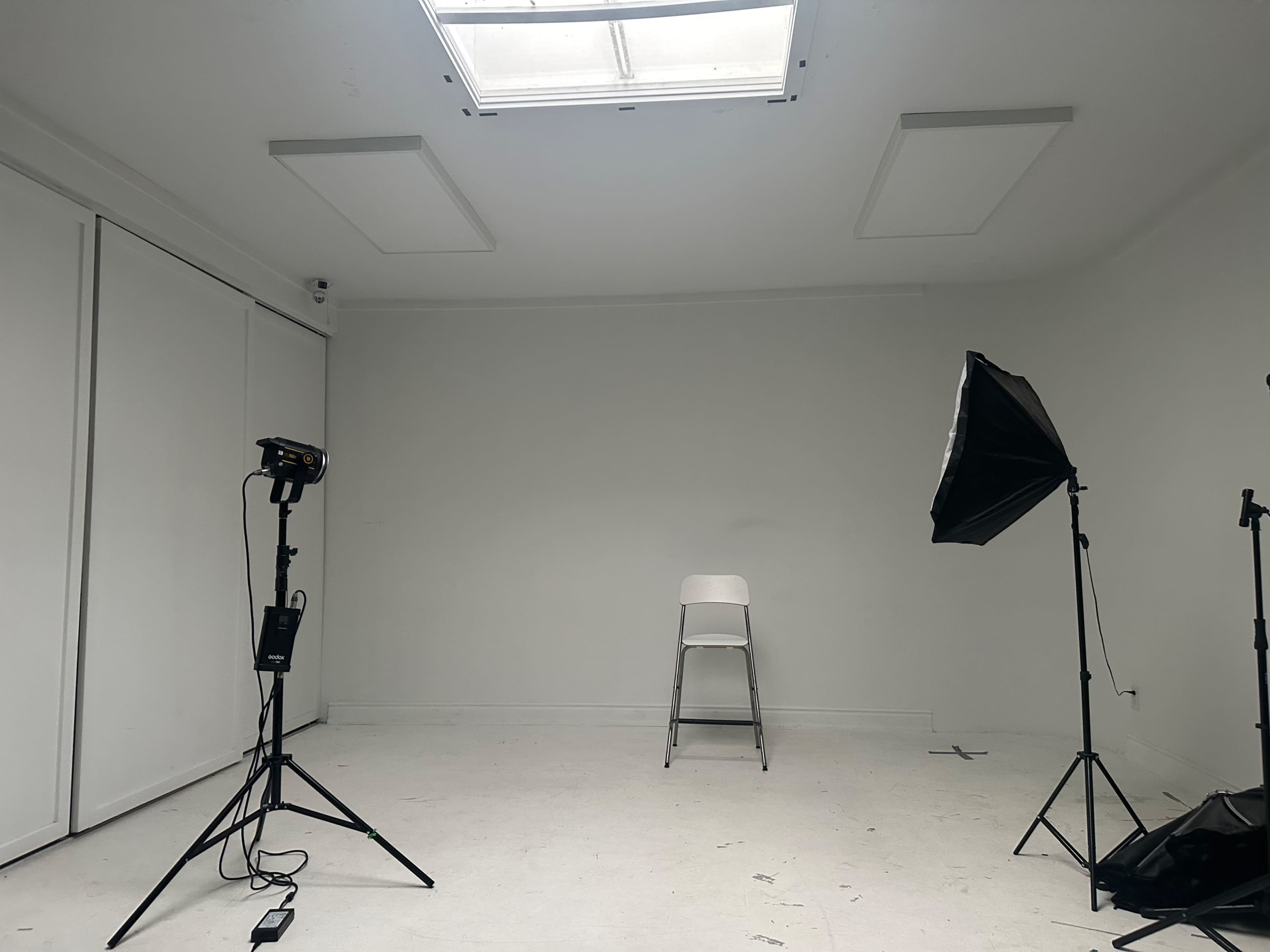 The image shows a minimalistic studio space with a white backdrop, a single chair in the center, and professional lighting equipment positioned to the side.