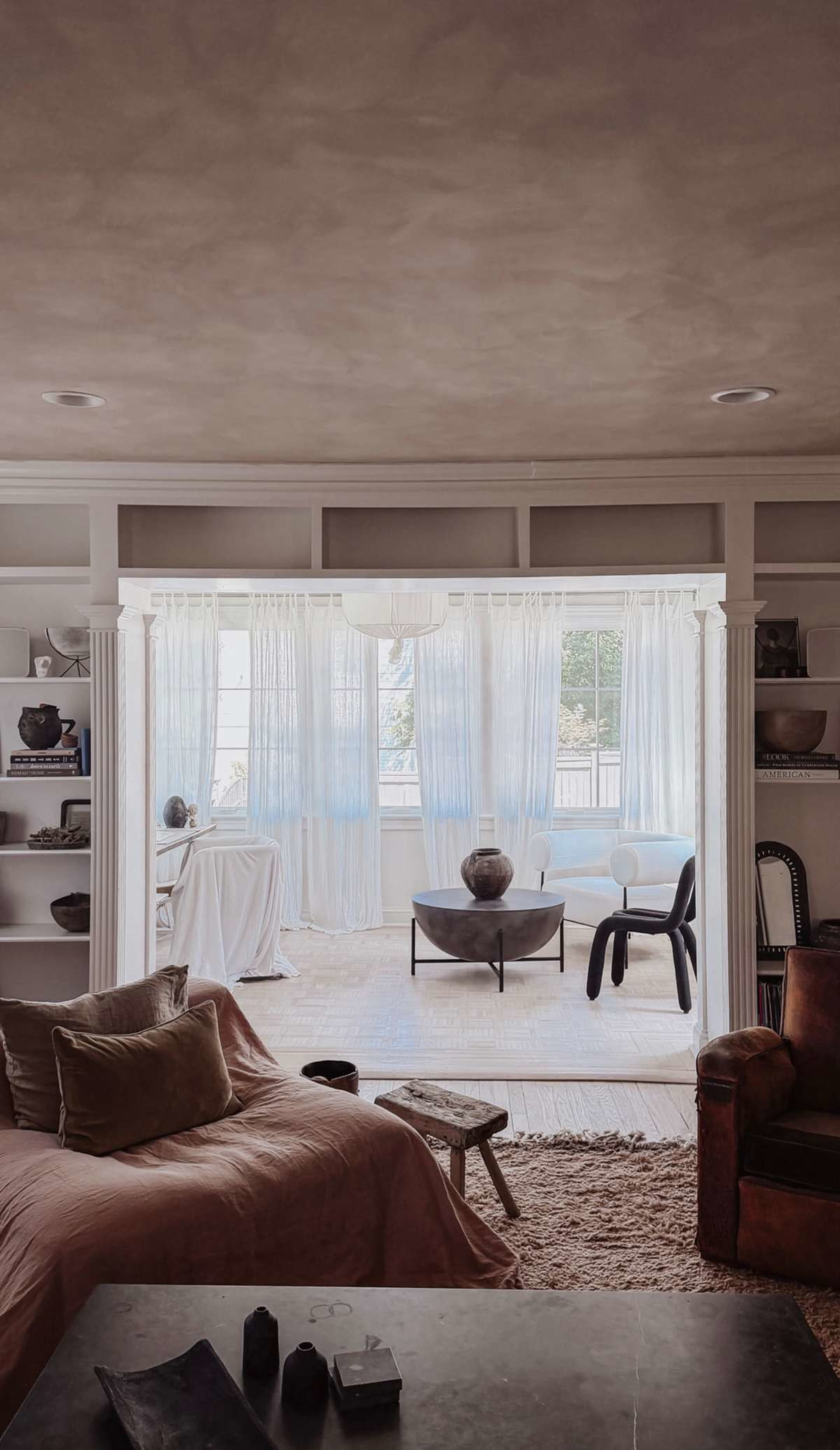 The image shows a spacious living room with a cozy sofa, decorative shelves, and large windows draped with sheer curtains allowing light to filter in.