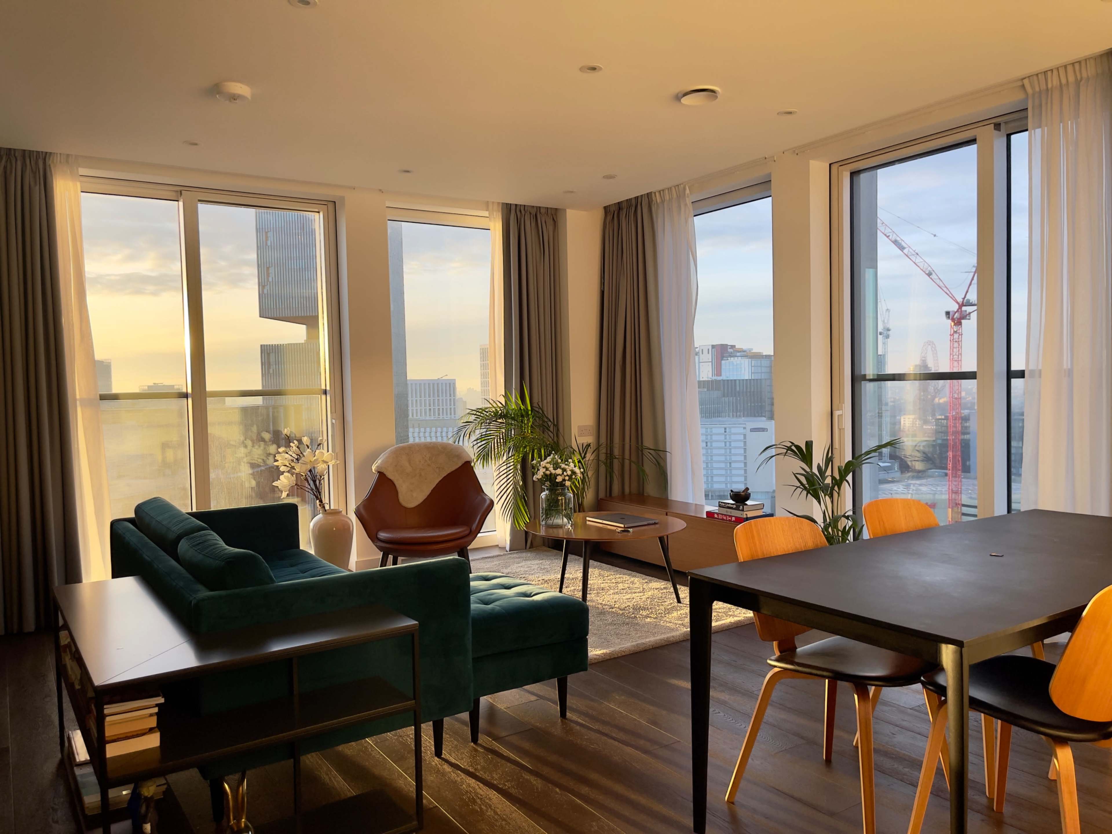 The image depicts a modern living room with large windows, a green couch, a wooden dining table, and a view of a cityscape at sunset.