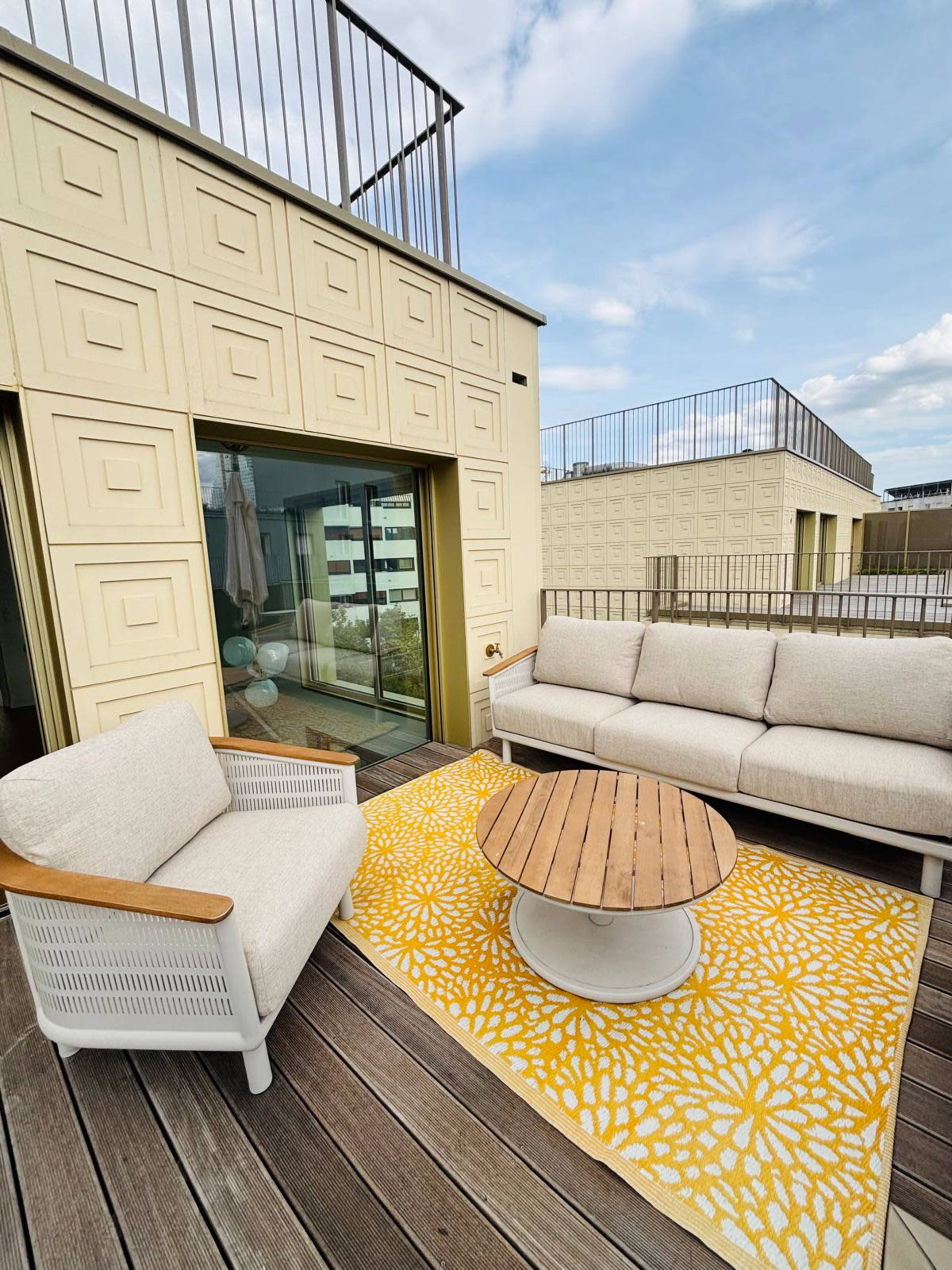 A rooftop terrace features a light-colored sofa, a beige armchair, and a round wooden coffee table on a yellow-patterned rug.