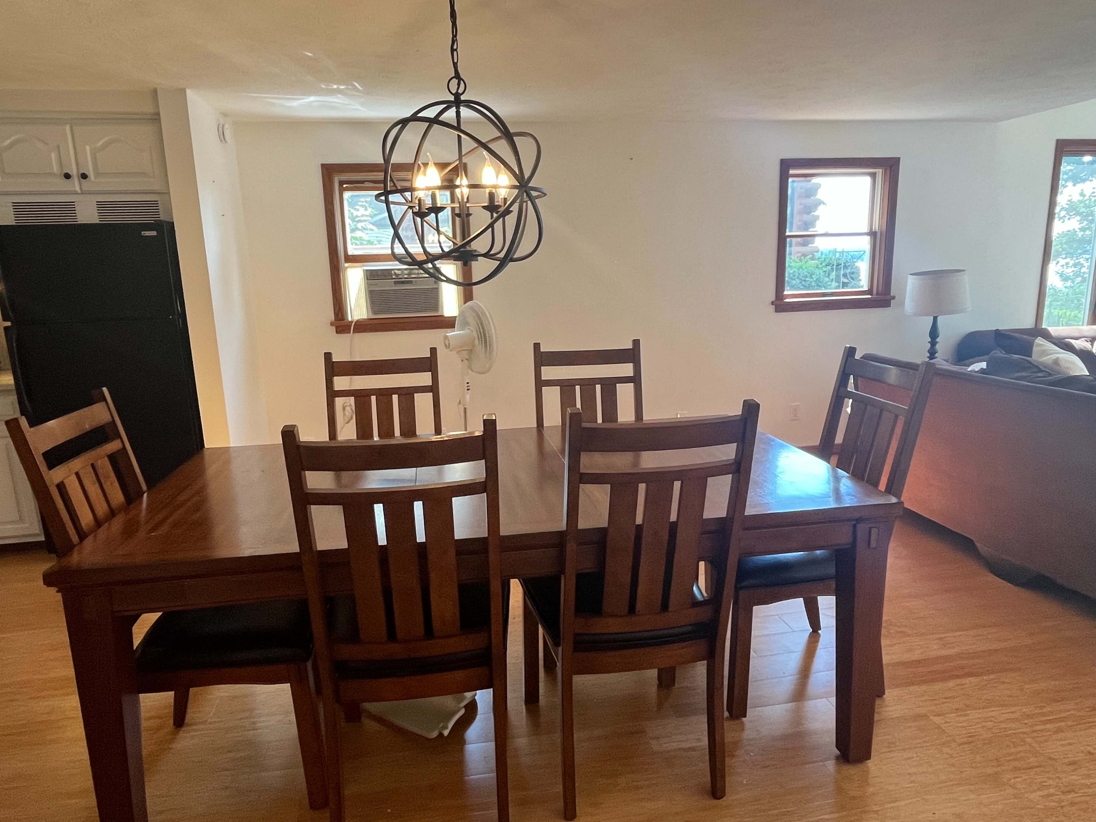 The image shows a dining area with a wooden table surrounded by six chairs, a black refrigerator in the background, and a living room area with a lamp and couch.