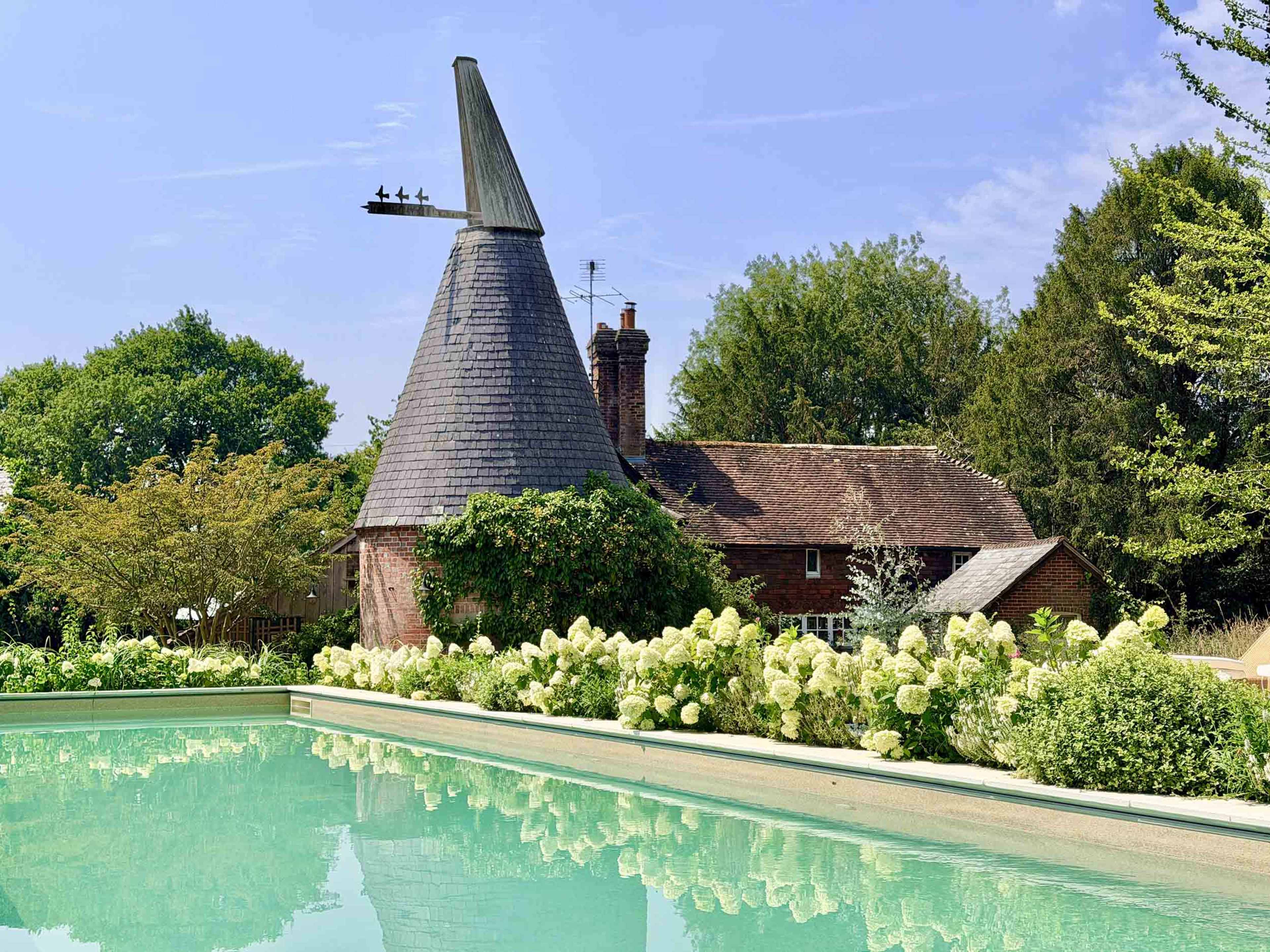 A traditional oast house with a conical roof is situated near a clear swimming pool, surrounded by blooming hydrangeas and lush greenery.