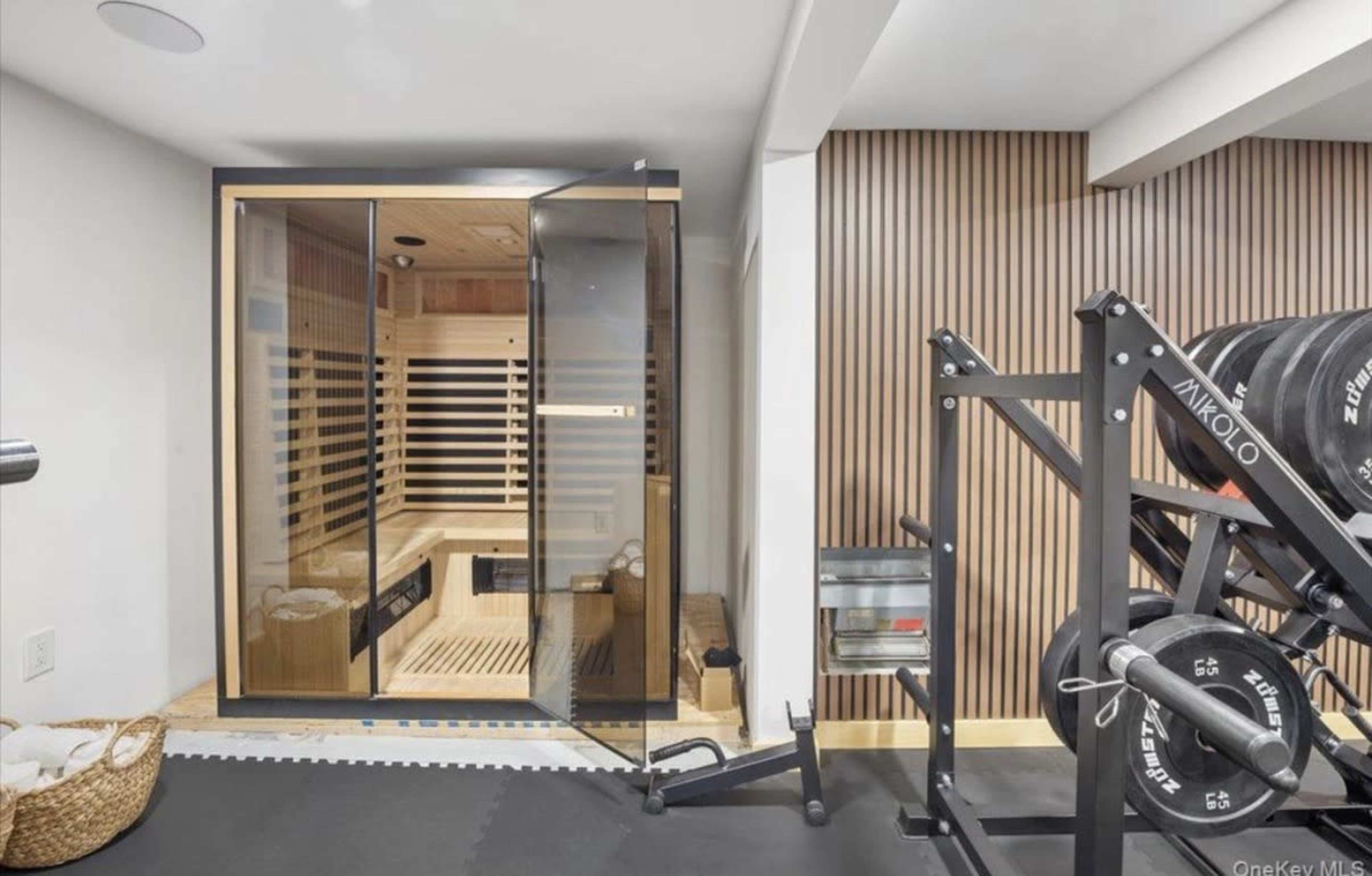The image shows a home gym featuring a glass-front sauna beside a weightlifting rack with free weights.