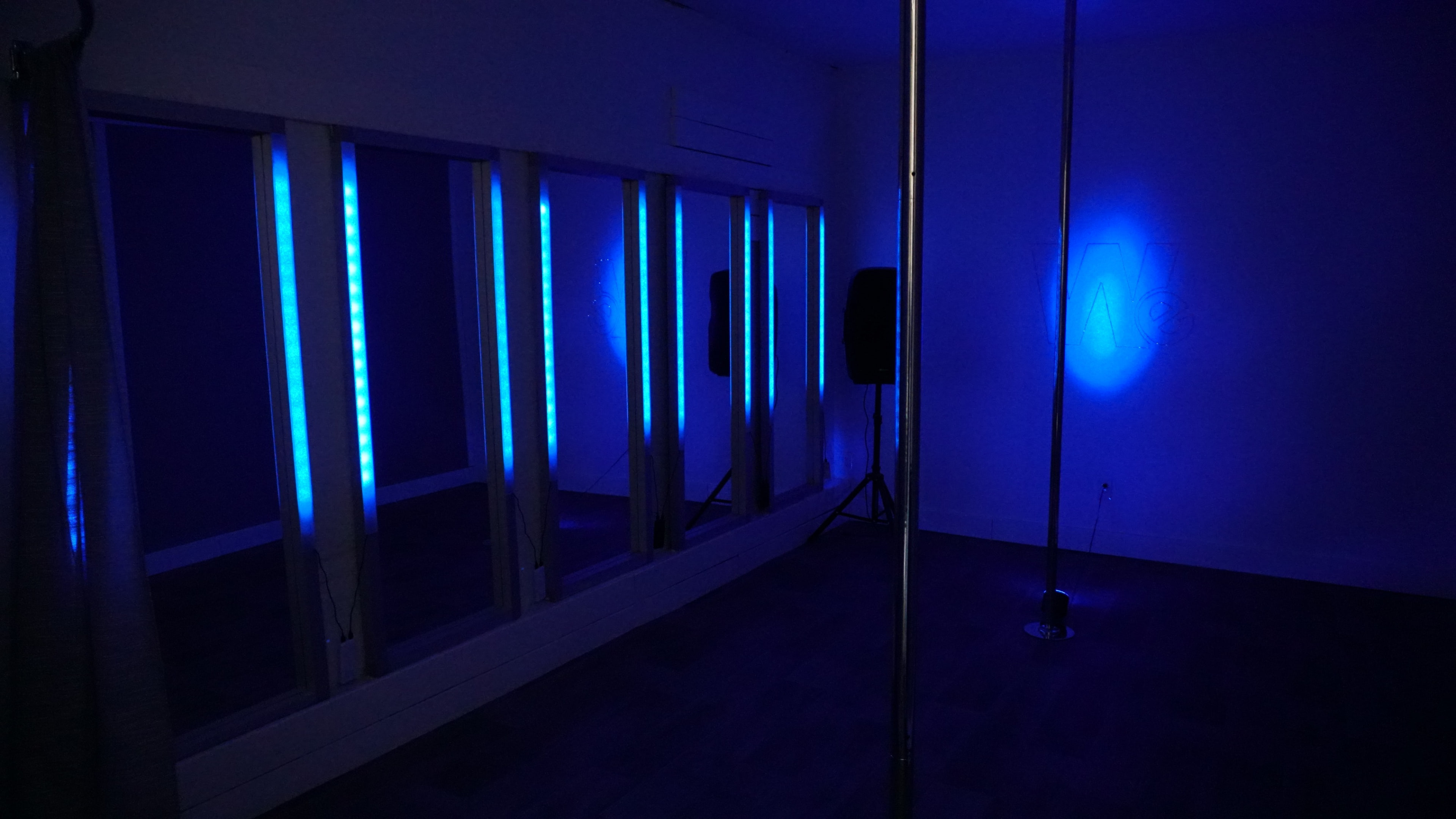A sparsely furnished room with blue ambient lighting, a mirror-lined wall, and a pole installed in the center.