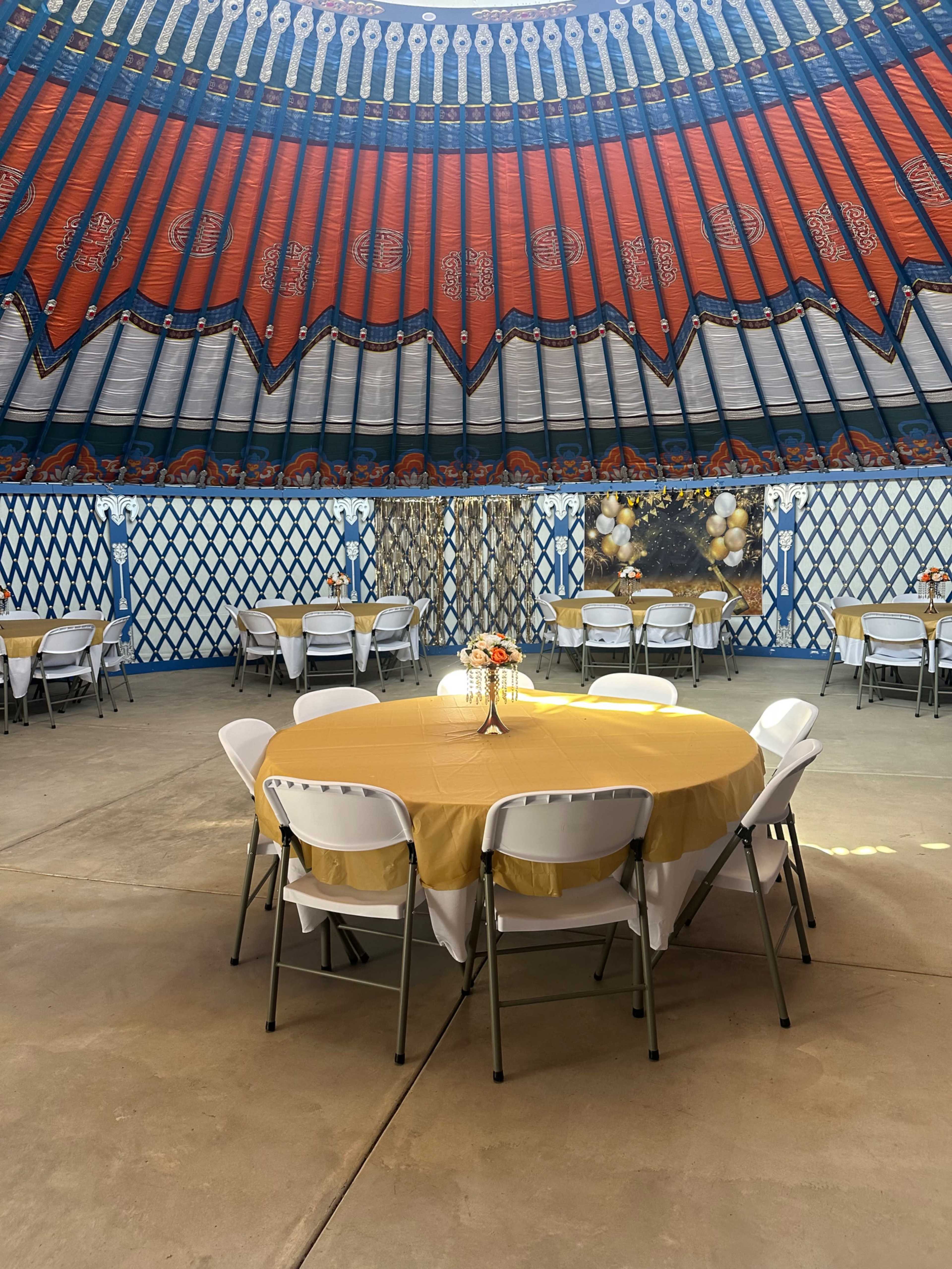 A round table with a yellow tablecloth and a centerpiece is set up inside a yurt with vibrant red and blue decor on the ceiling.