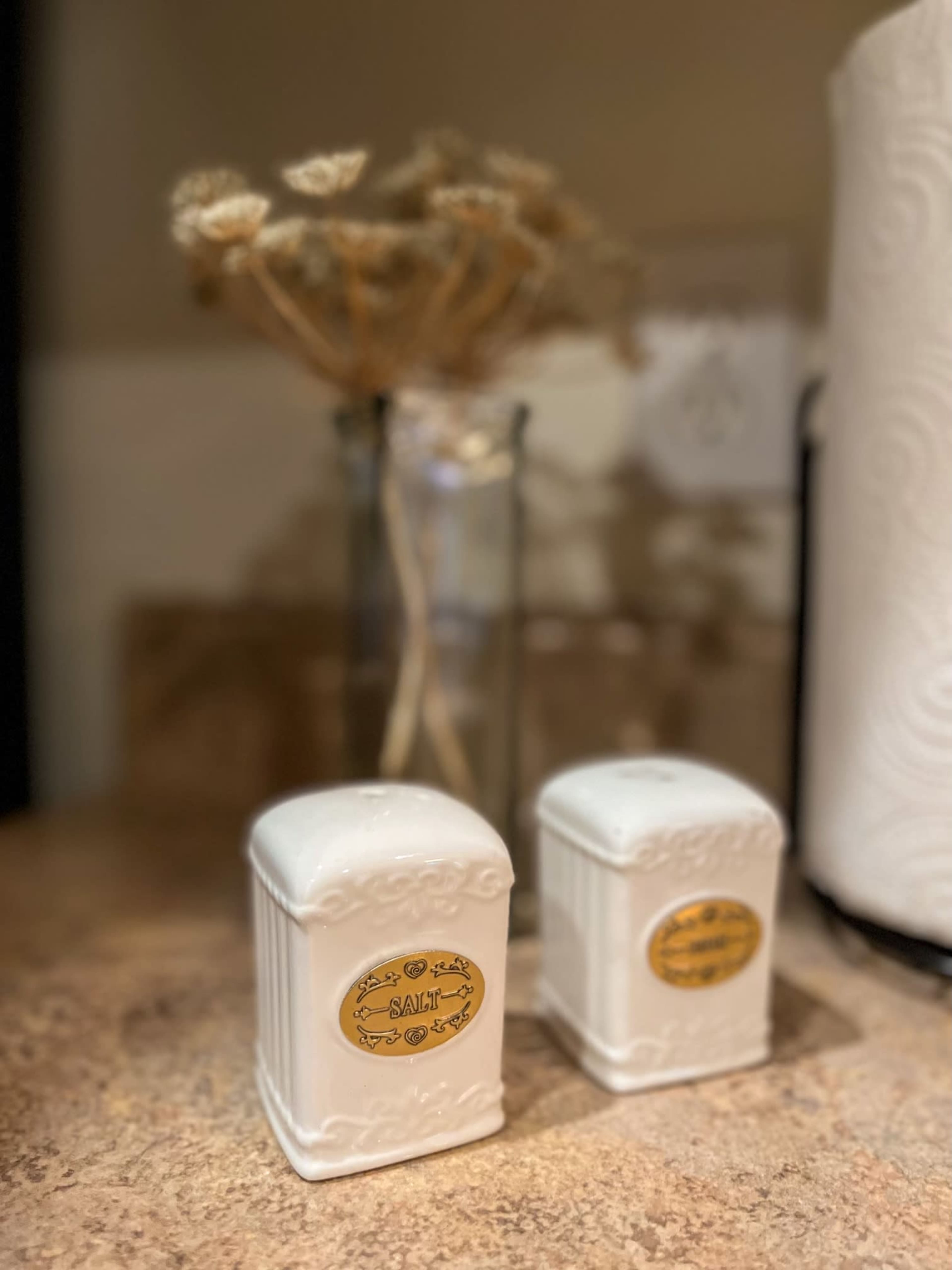 Two white salt shakers with gold labels on a kitchen countertop beside a glass vase containing dried flowers.