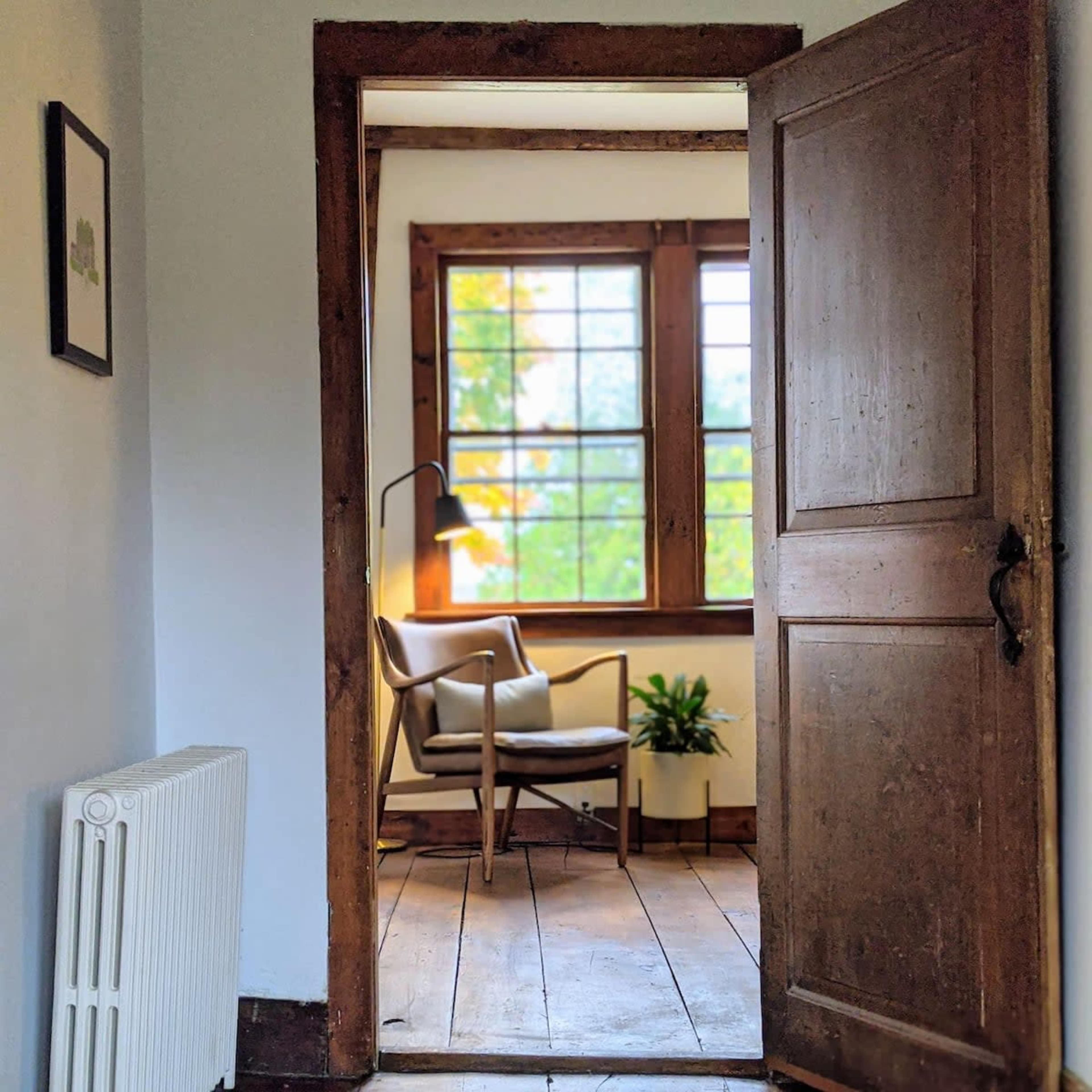 A wooden door opens to a well-lit room featuring a chair and a lamp near a window with a view of greenery.