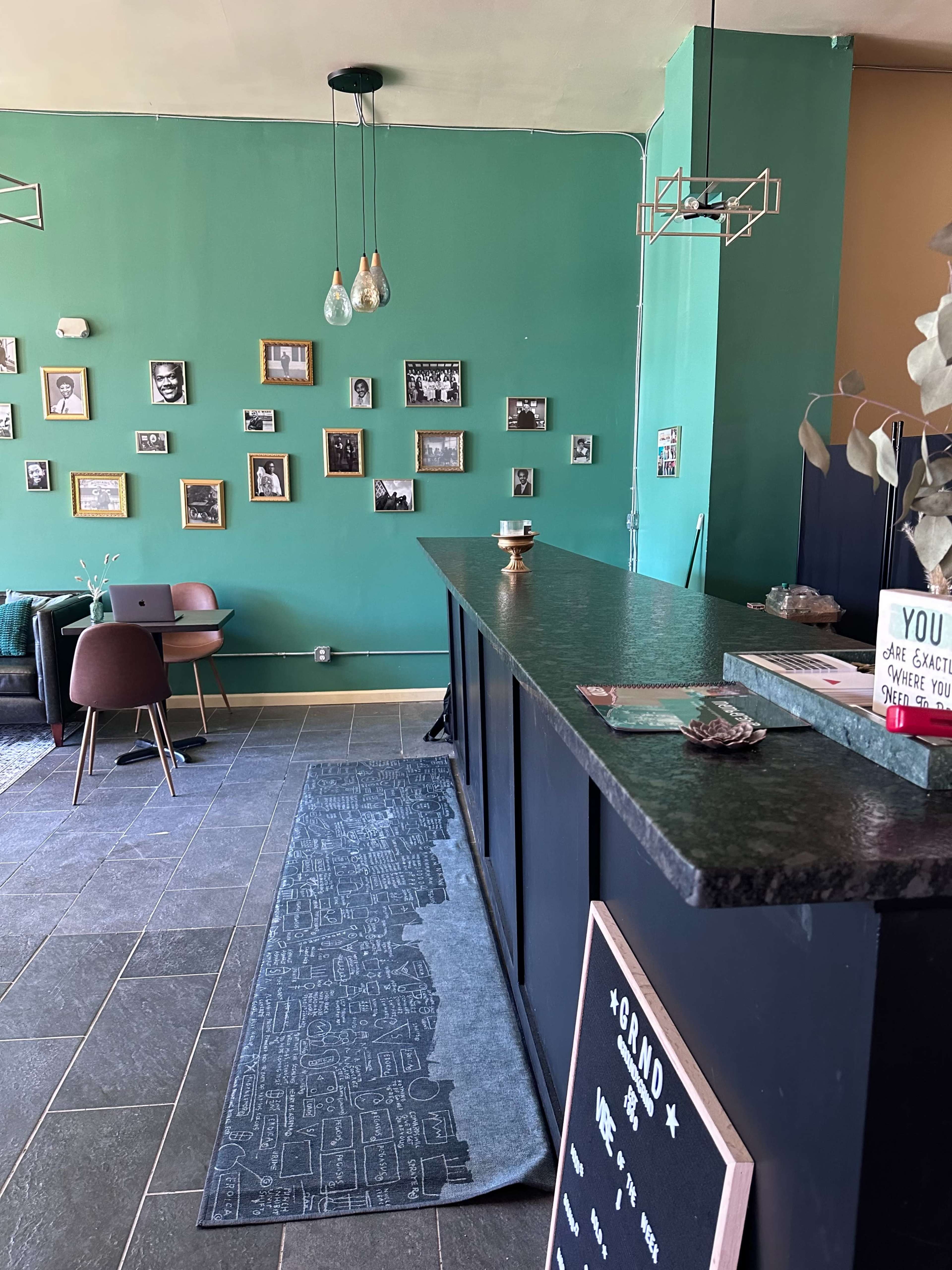 A modern cafe interior with a dark countertop, green walls adorned with framed photos, and seating arrangements on one side.