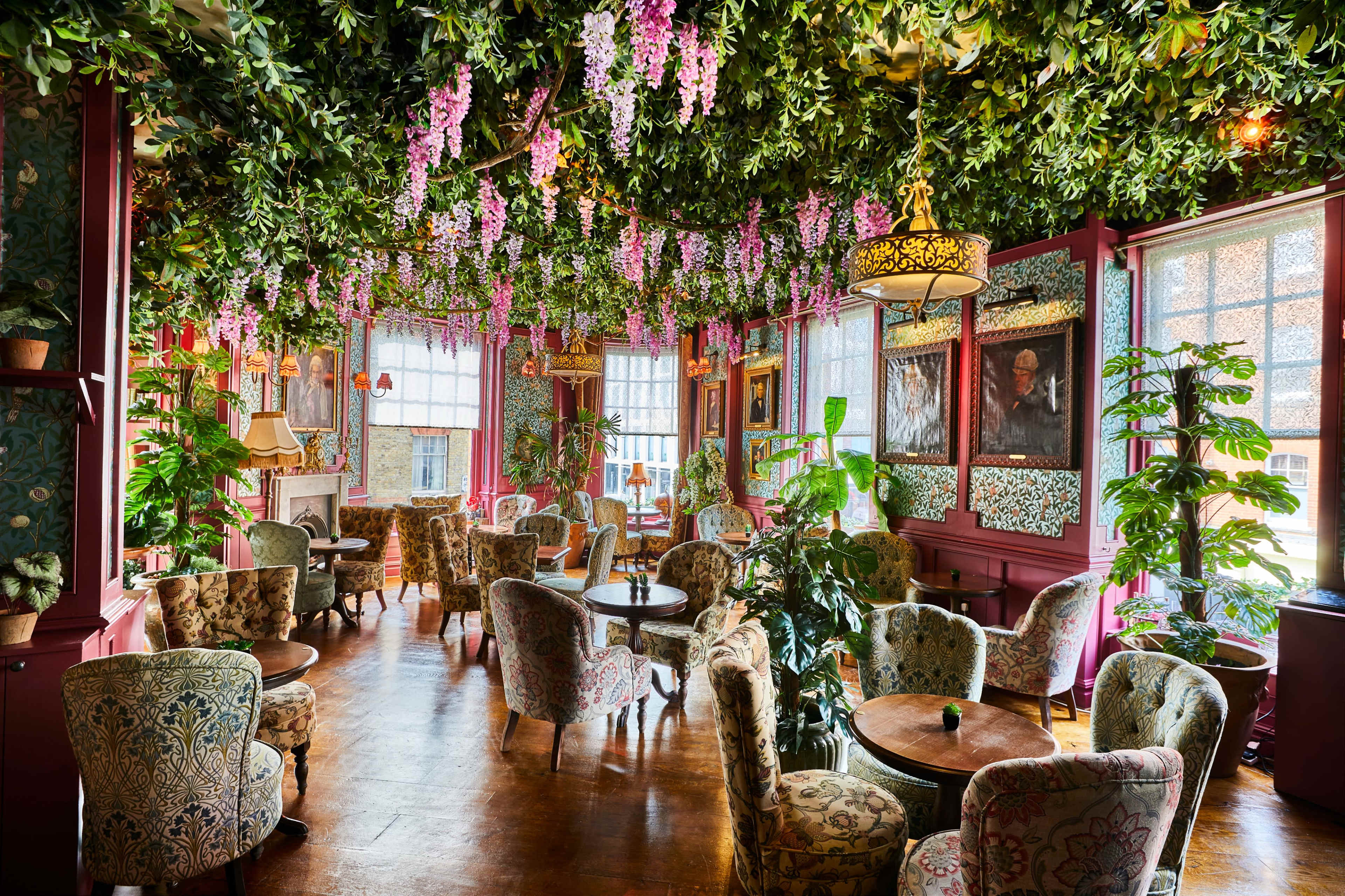 The image shows a vibrant café with floral decorations hanging from the ceiling, featuring upholstered seating and numerous potted plants throughout the room.