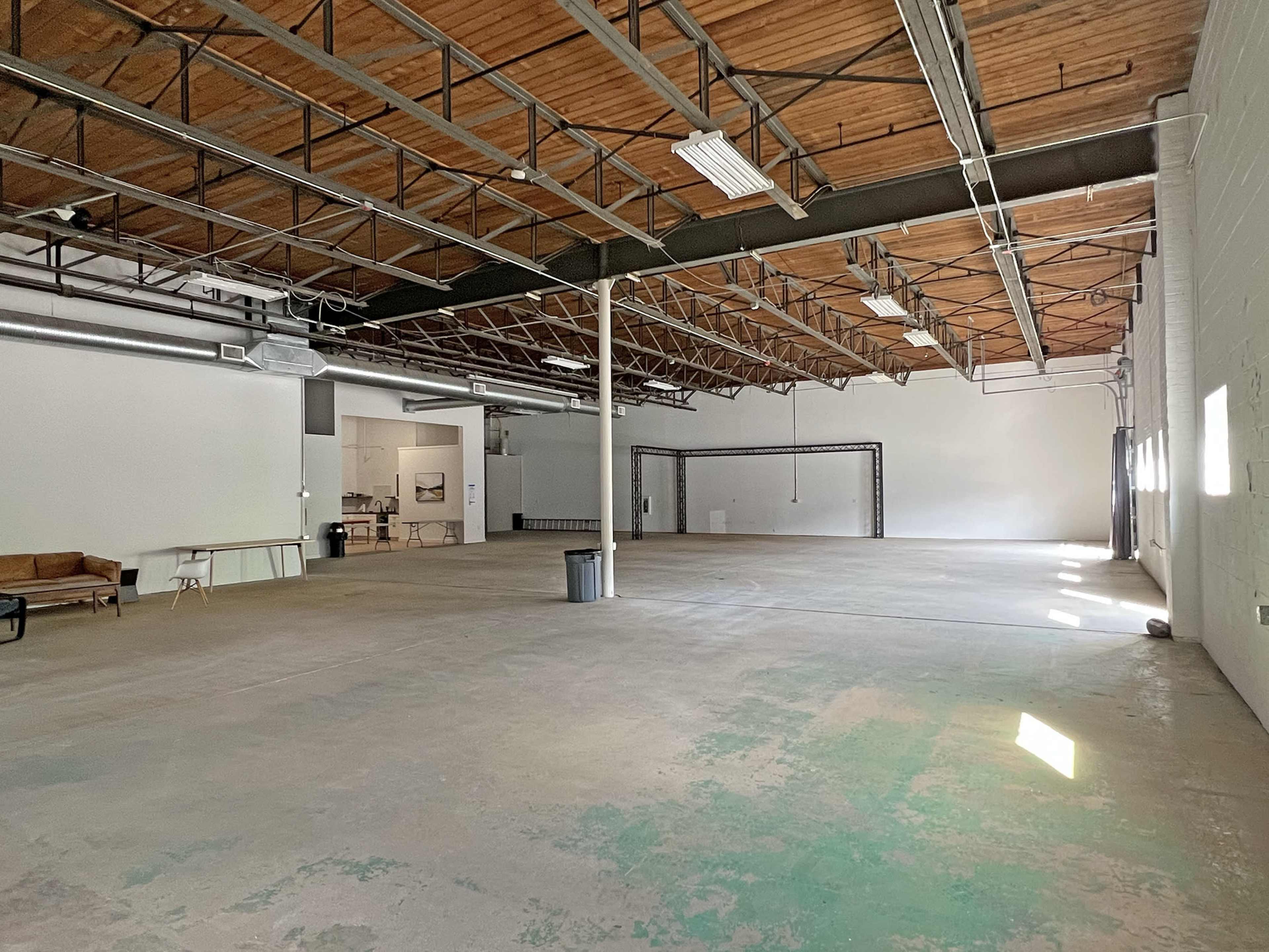 A large, empty warehouse space with exposed wooden beams, concrete flooring, and minimal furnishings.