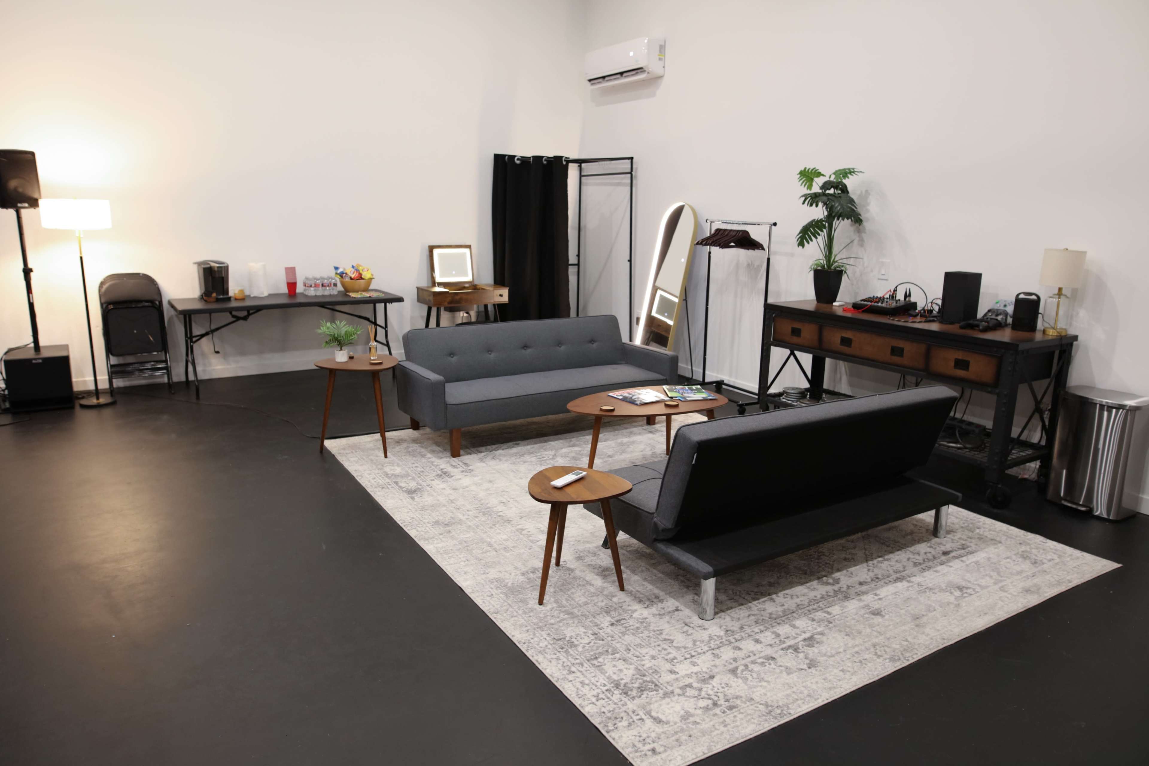 The image shows a modern, minimalistic interior space featuring two gray sofas, a rug, small wooden tables, a coffee station, and a clothing rack against a plain backdrop.