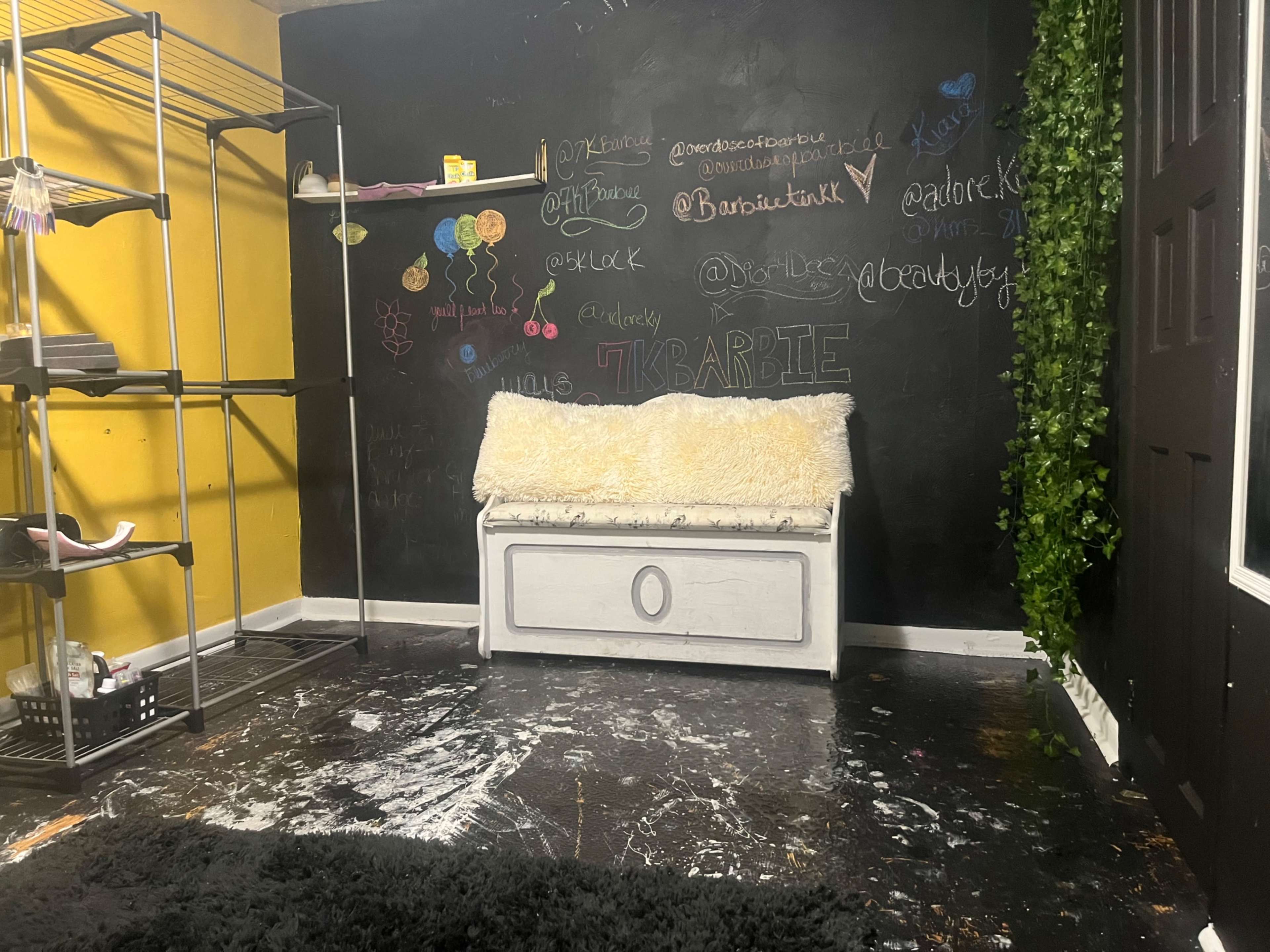 A dimly lit room with a black chalkboard wall covered in colorful chalk writings, a white fur bench in the center, and minimal furnishings.