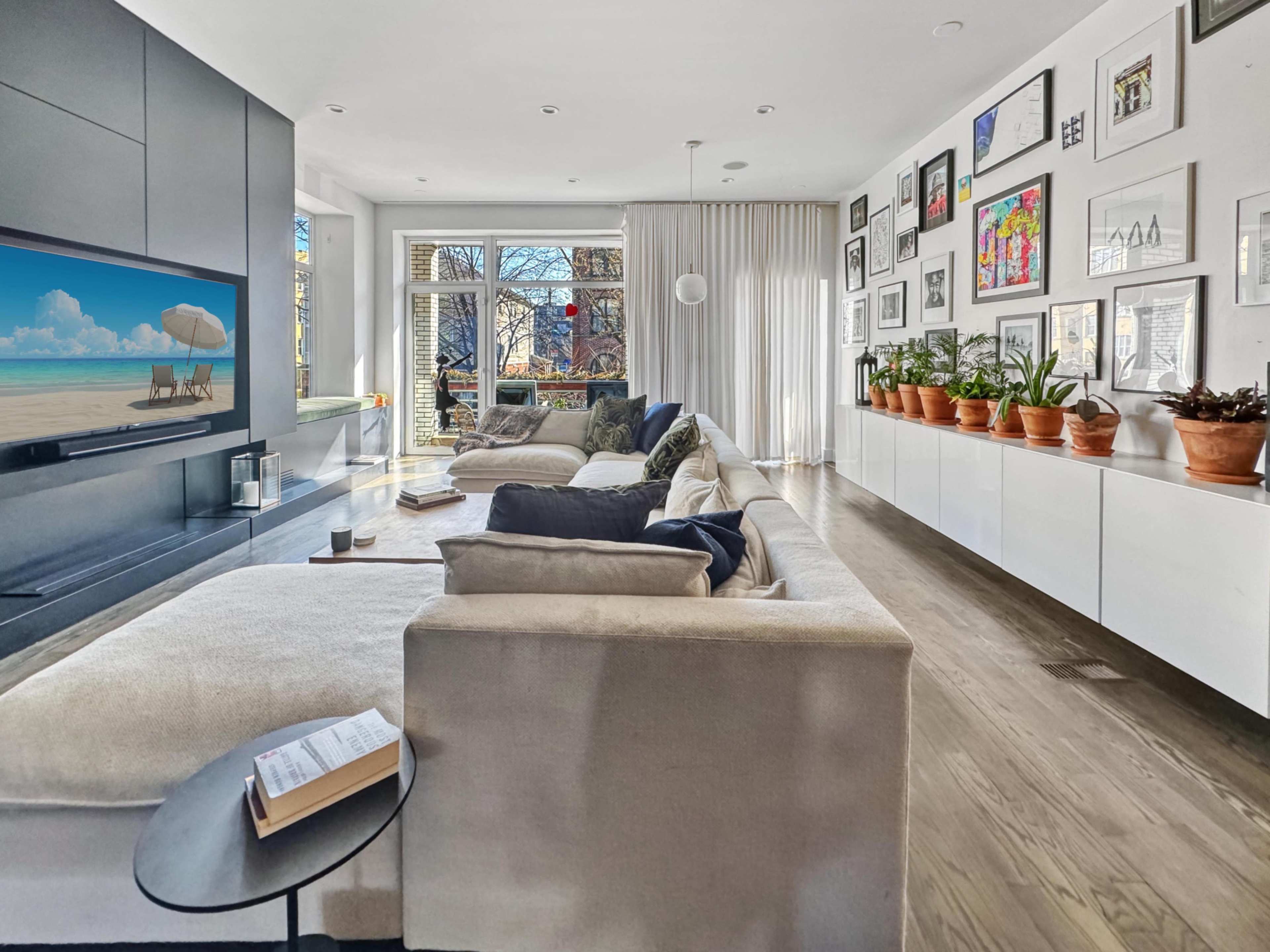 A modern living room features a large sectional sofa, a flat-screen TV mounted on a wall, and a gallery of framed artwork alongside potted plants on a sleek sideboard.