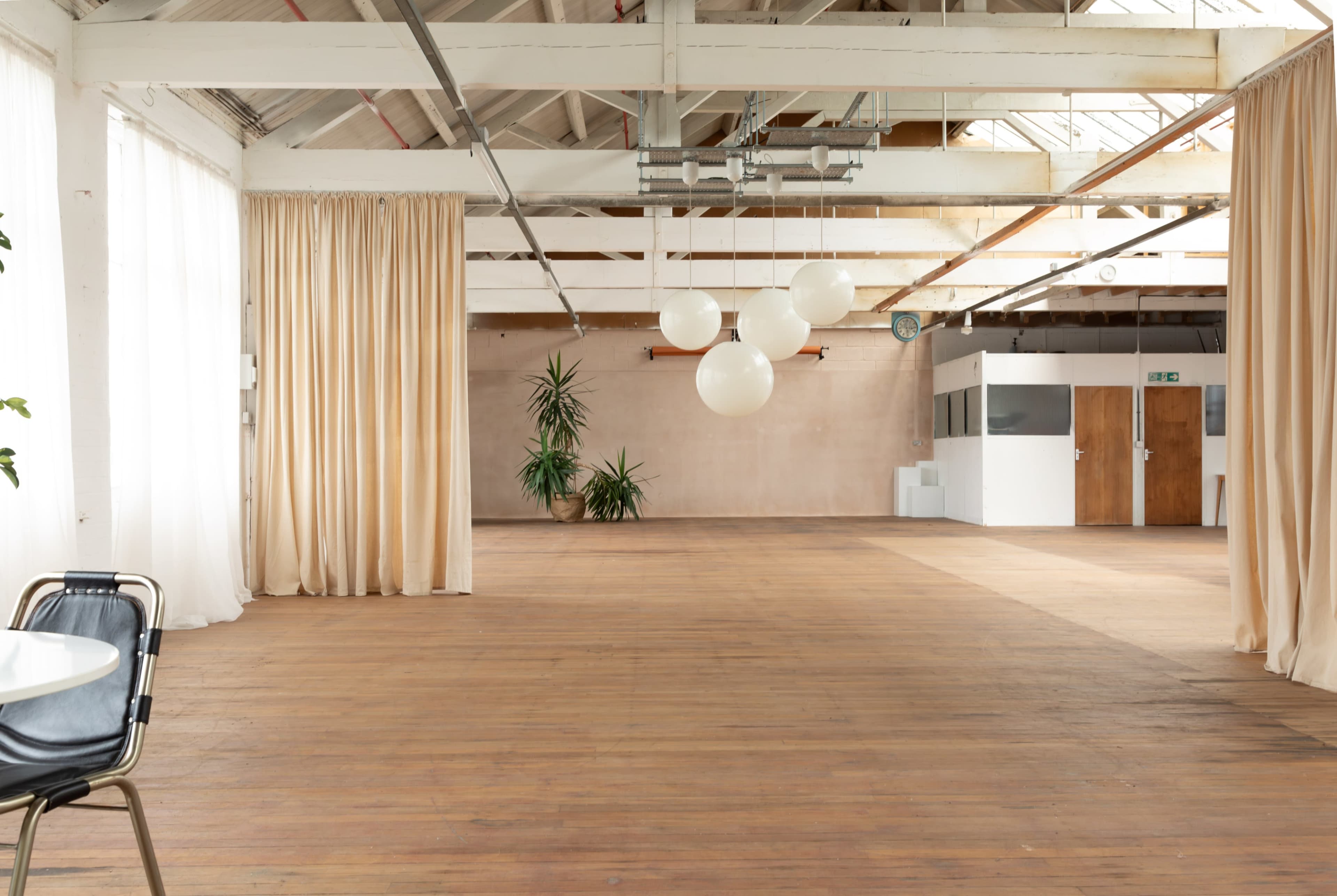 The image shows an empty, spacious interior with wooden flooring, high ceilings, large windows covered in sheer curtains, and a few plants.