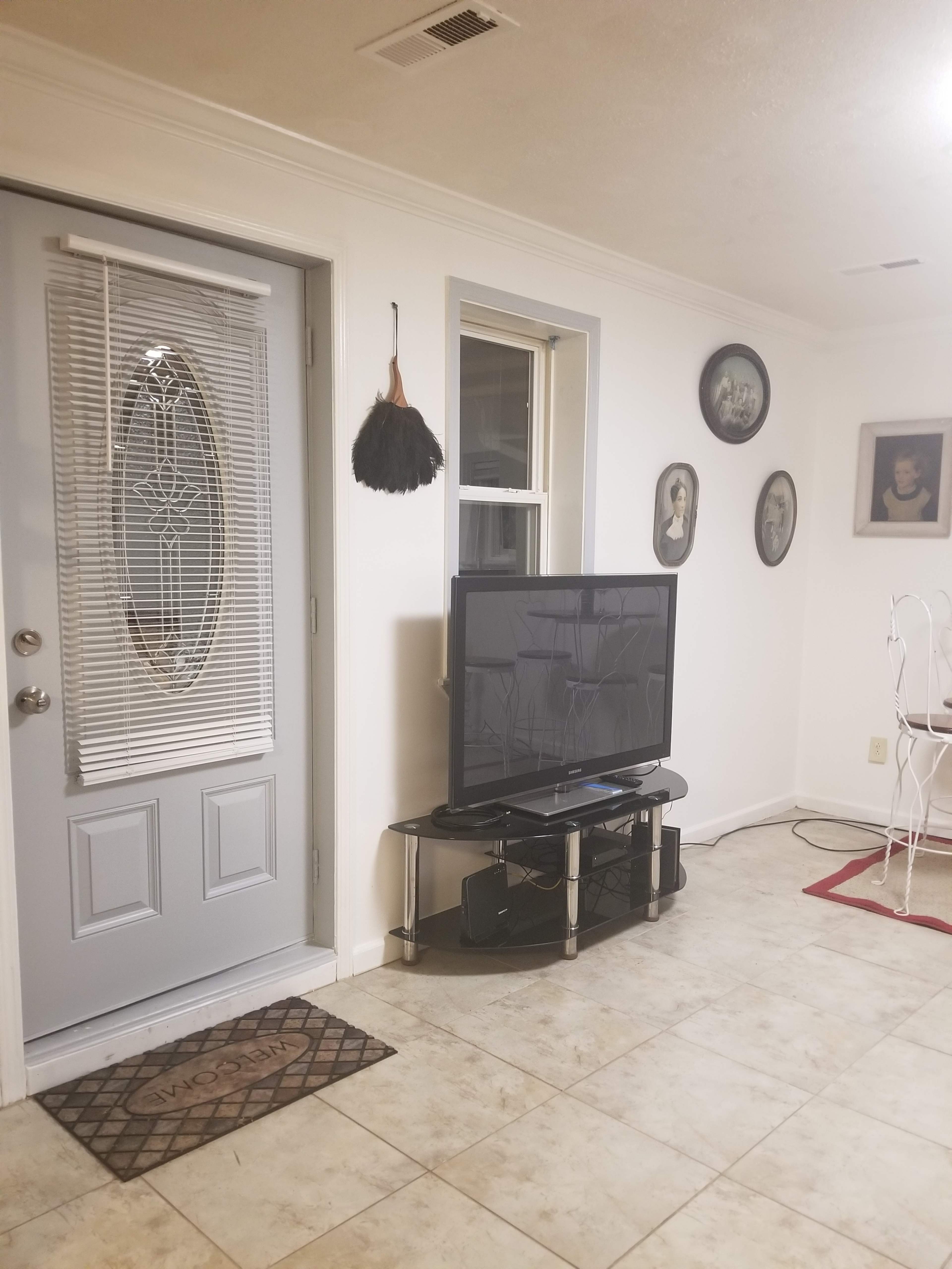A living room with a front door, a television on a stand, and framed images on the wall.