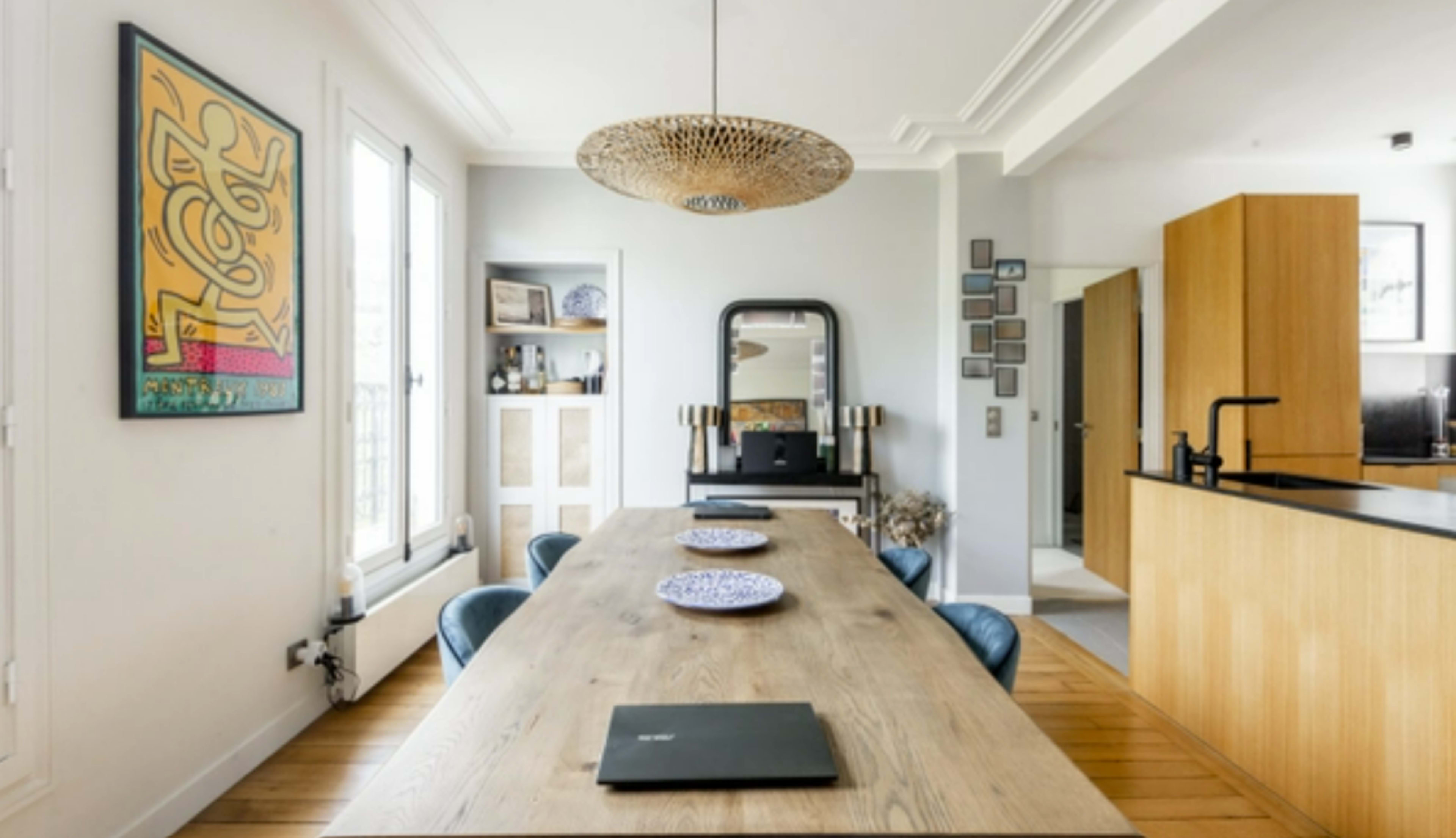 A spacious dining area features a long wooden table, four chairs, a decorative mirror, and a modern pendant light, with a kitchen visible in the background.