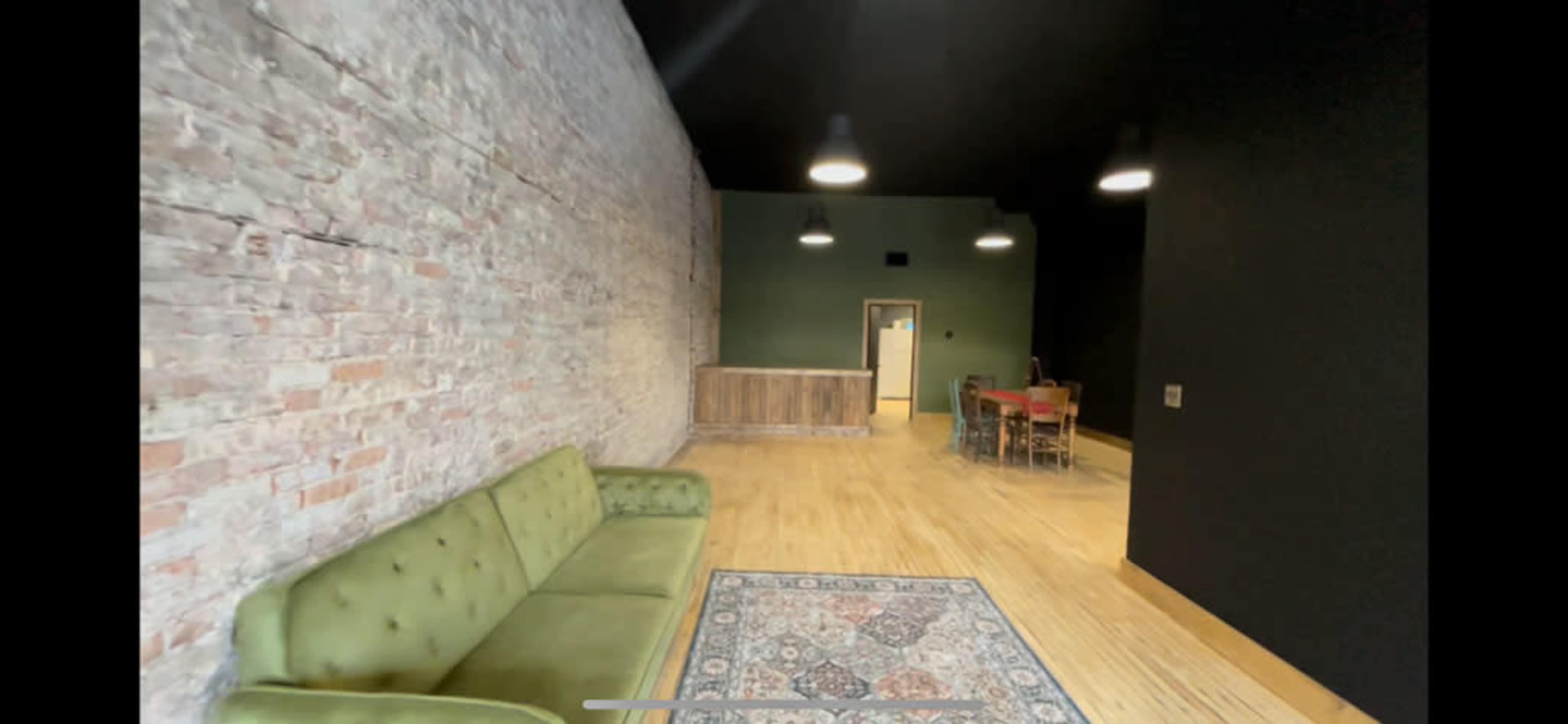 A spacious interior with a green velvet sofa, a patterned rug, and a wooden counter at the back against a brick and black wall.