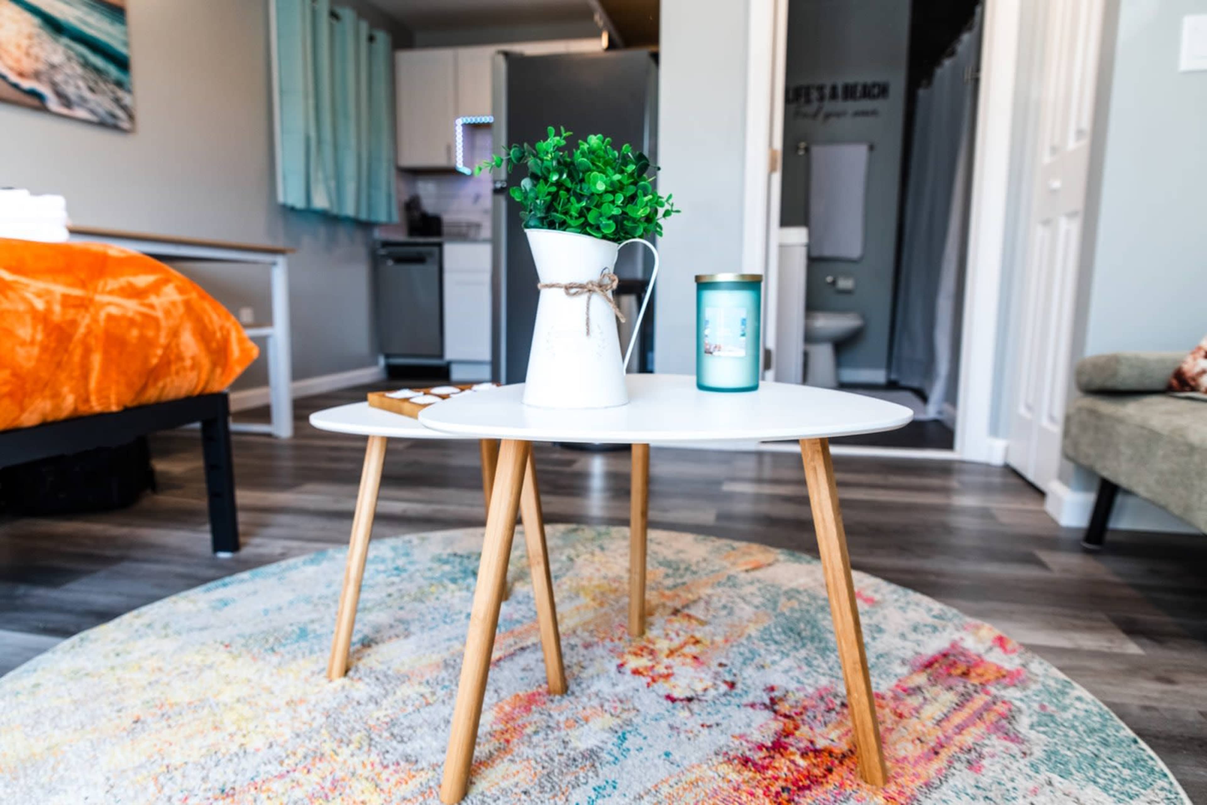A round coffee table with a plant and a candle sits on a colorful rug in a small living space.