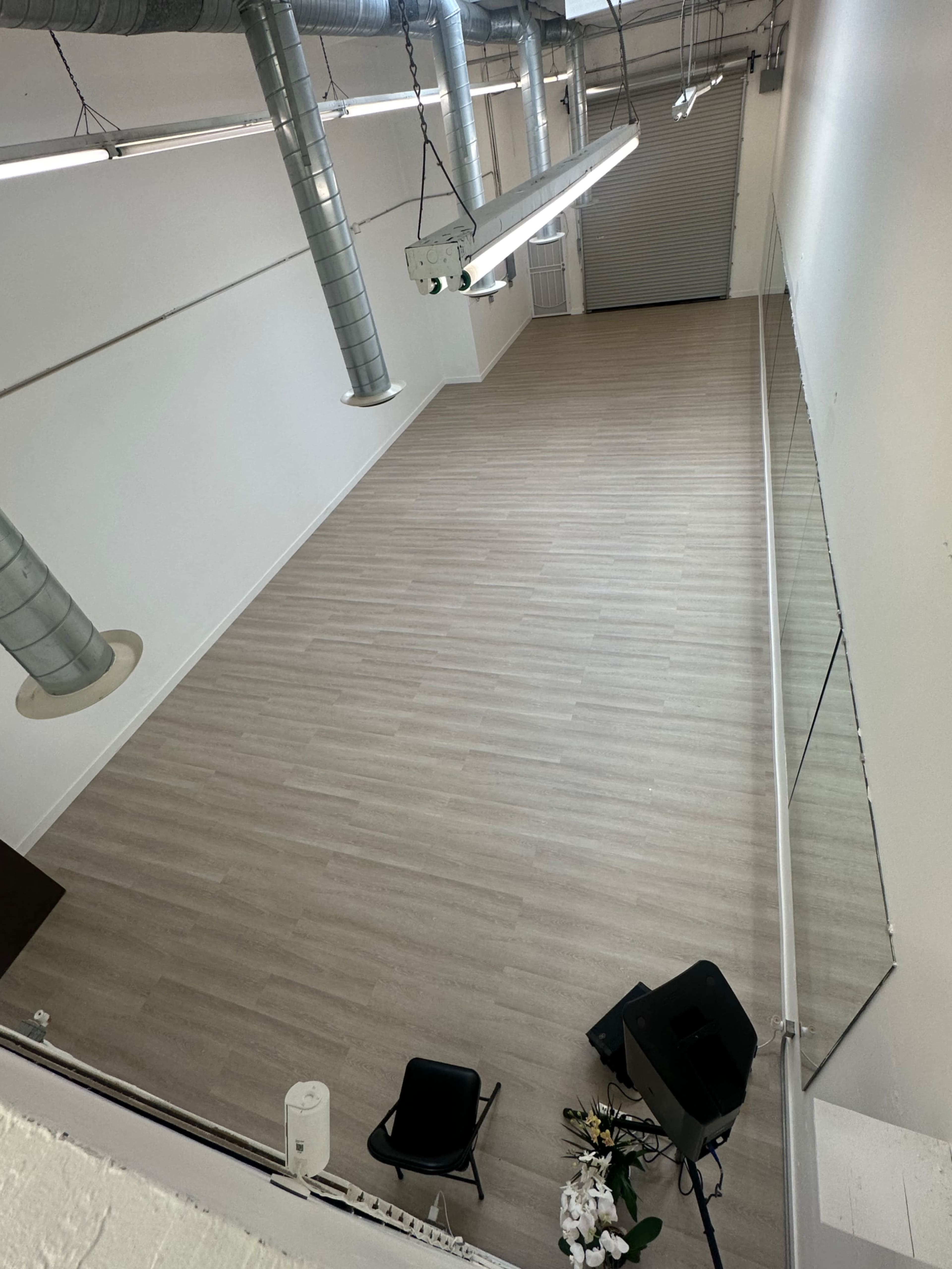 The image shows a spacious, empty interior room with light-colored wooden flooring, high ceilings, and large mirrors along one wall.