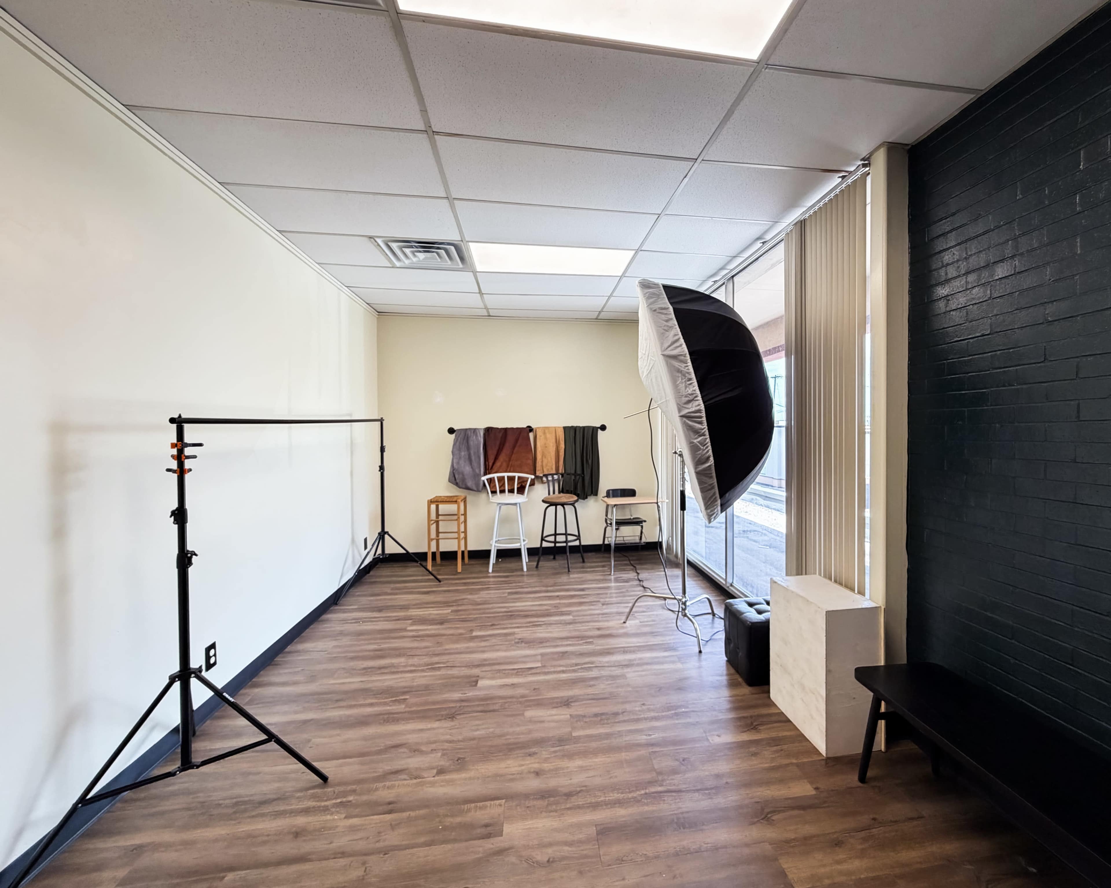 The image shows a vacant photography studio with a plain wall, stands for lighting equipment, a few stools, and various hanging fabrics.