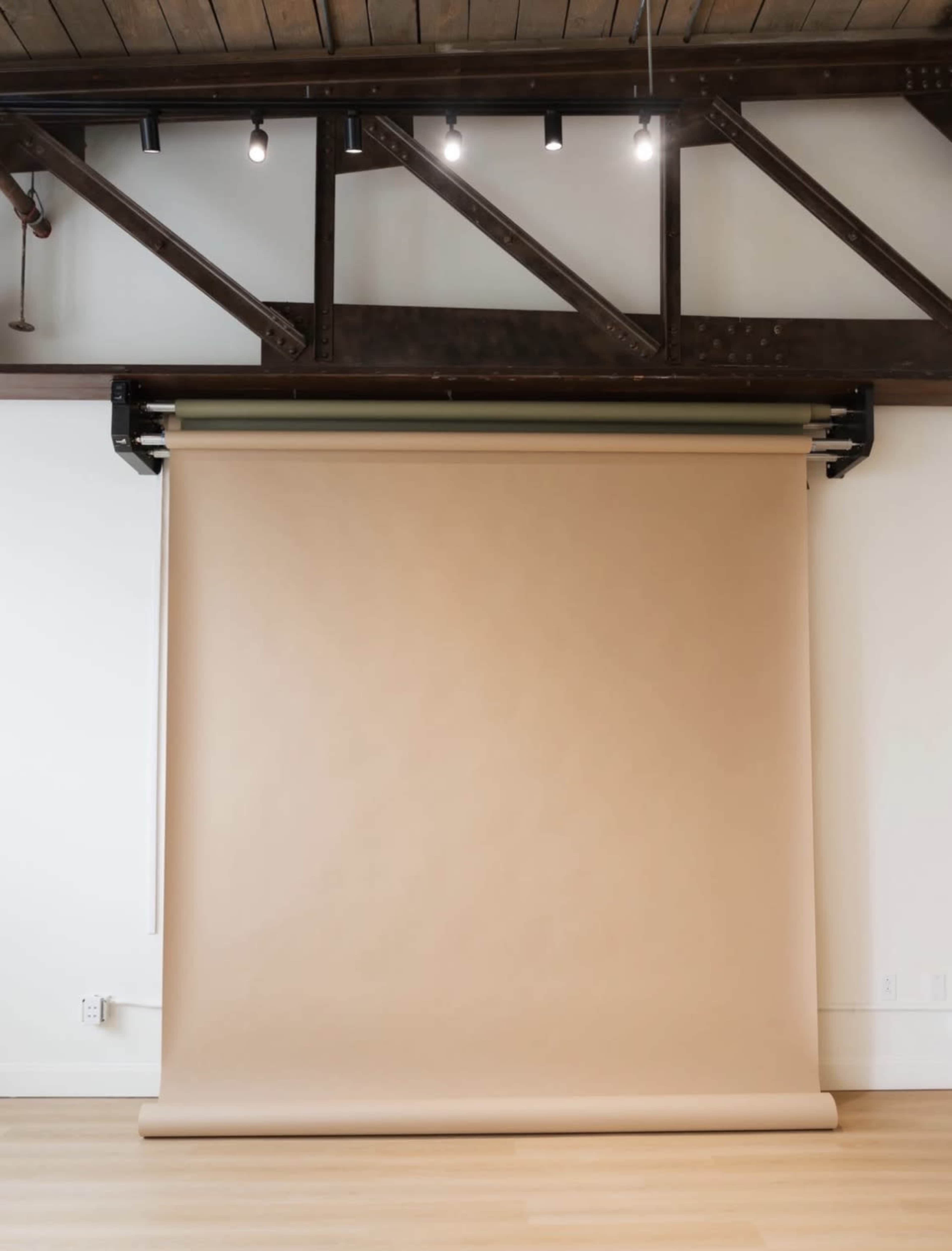 A large roll of beige backdrop paper is suspended from a black mounting system against a white wall in a room with wooden beams.