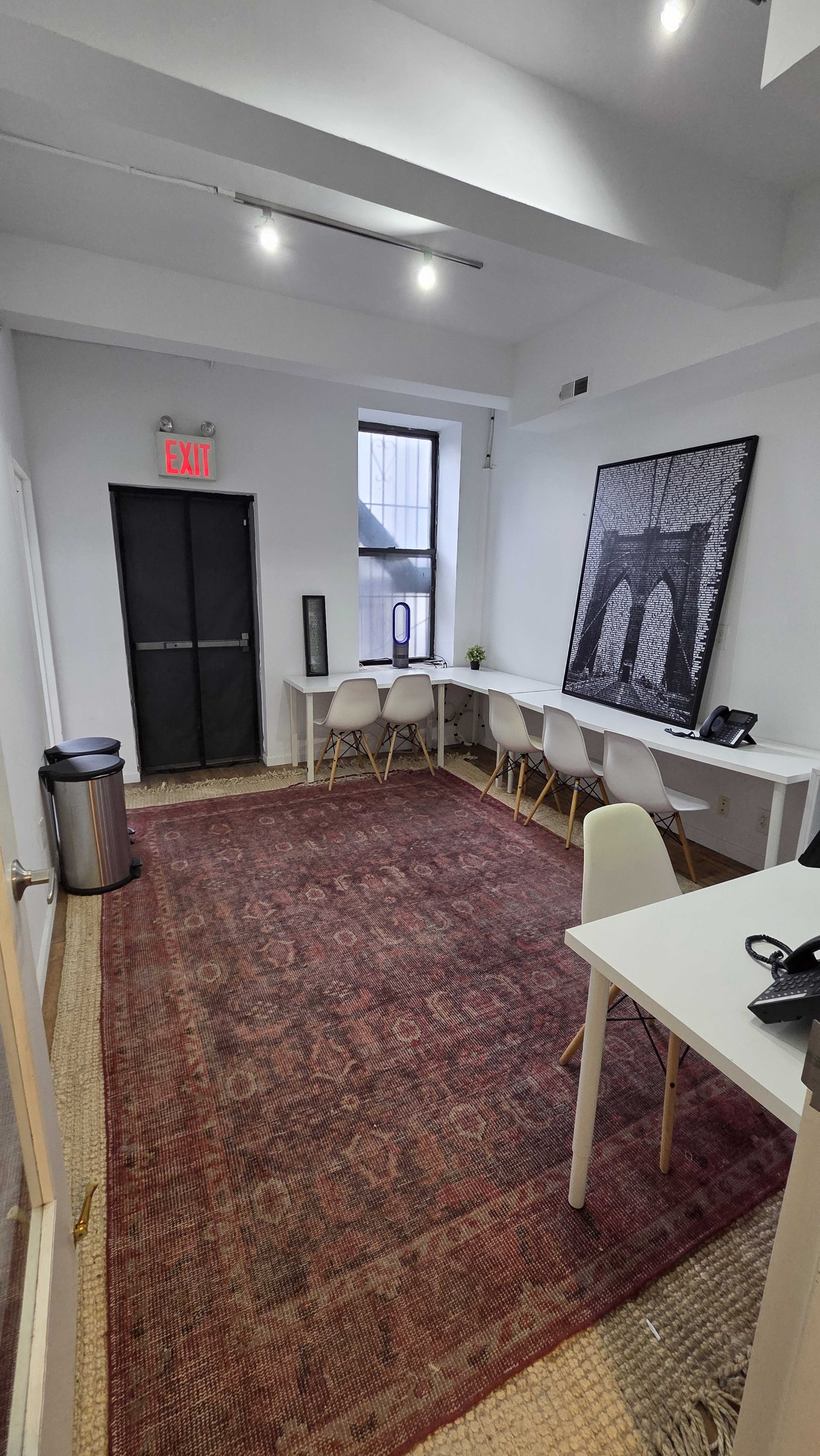 The image shows a modern office space with a large rug, a long white table with several chairs, a trash can, and a black-and-white photograph on the wall.