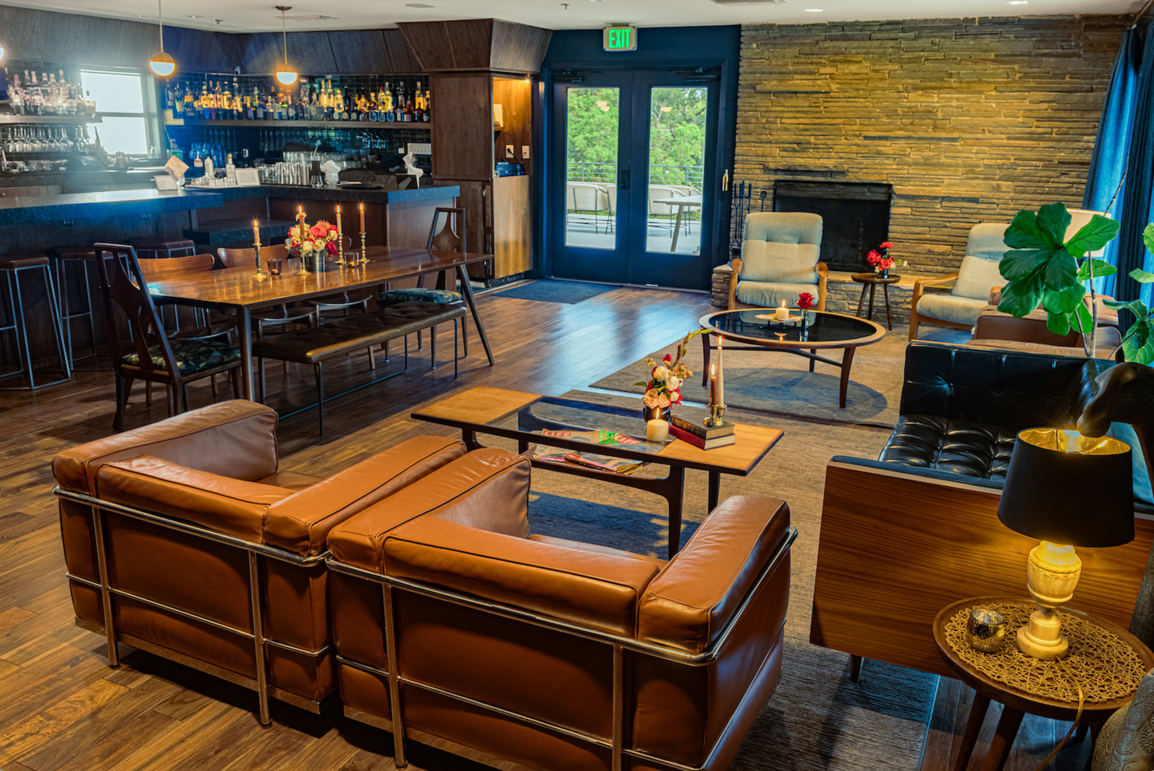 The image shows a modern lounge area featuring seating arrangements with brown leather couches and chairs, a wooden dining table, a bar in the background, and decorative plants.
