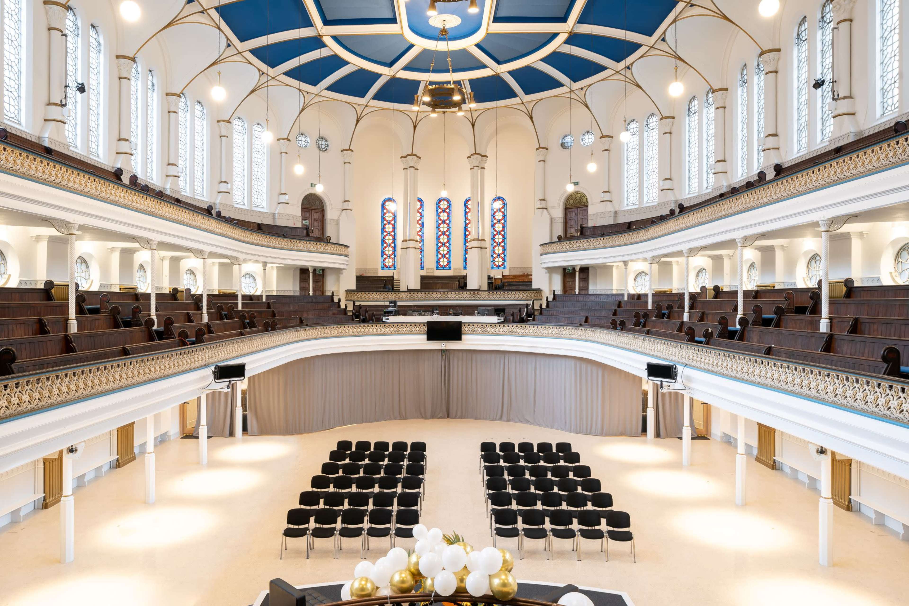 A spacious, elegantly designed auditorium with tiered seating arranged in front of a stage, featuring large windows and a decorative ceiling.