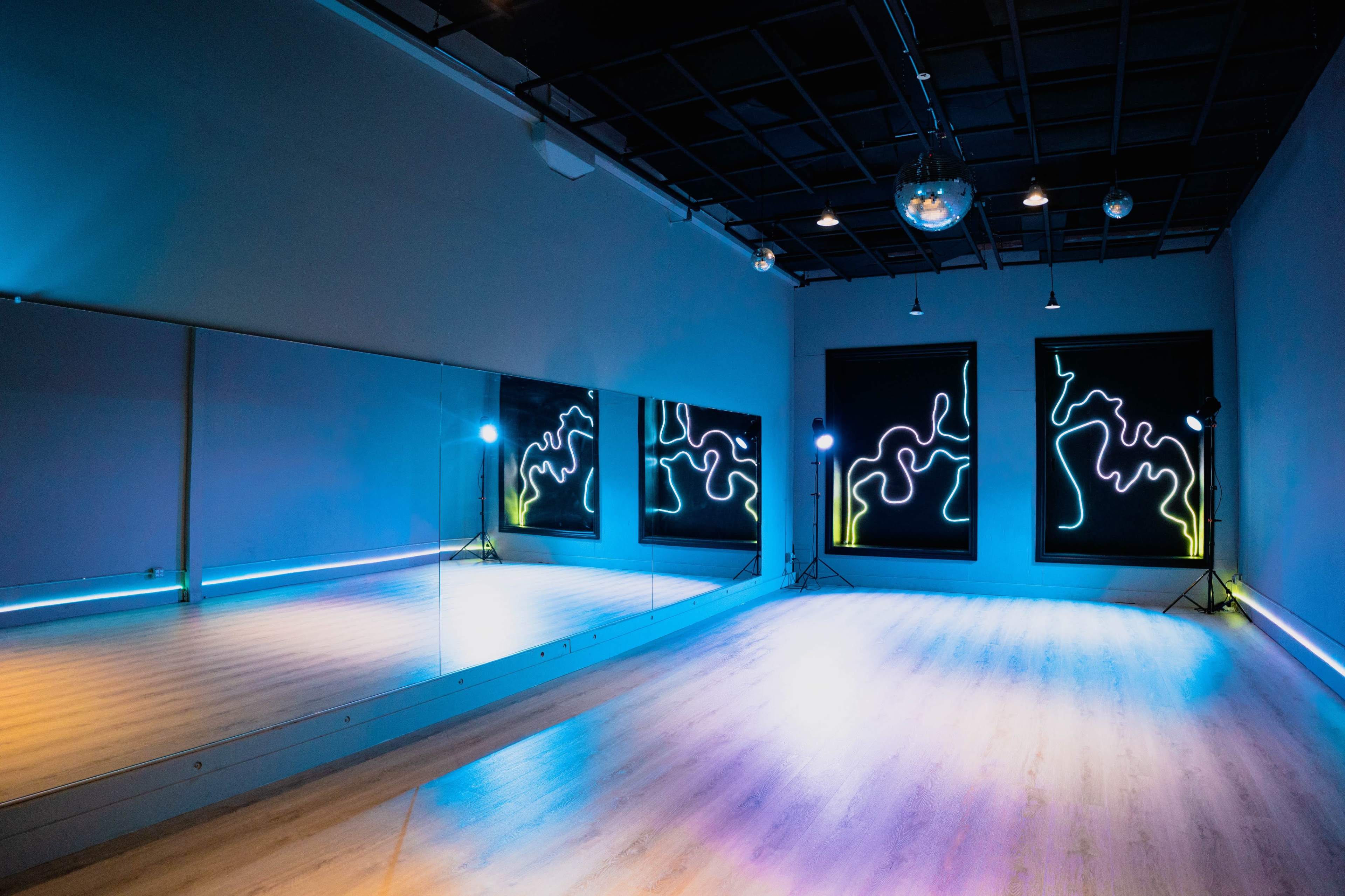 The image shows a spacious dance studio with large mirrors, colorful neon light decorations on the walls, and a wooden floor.