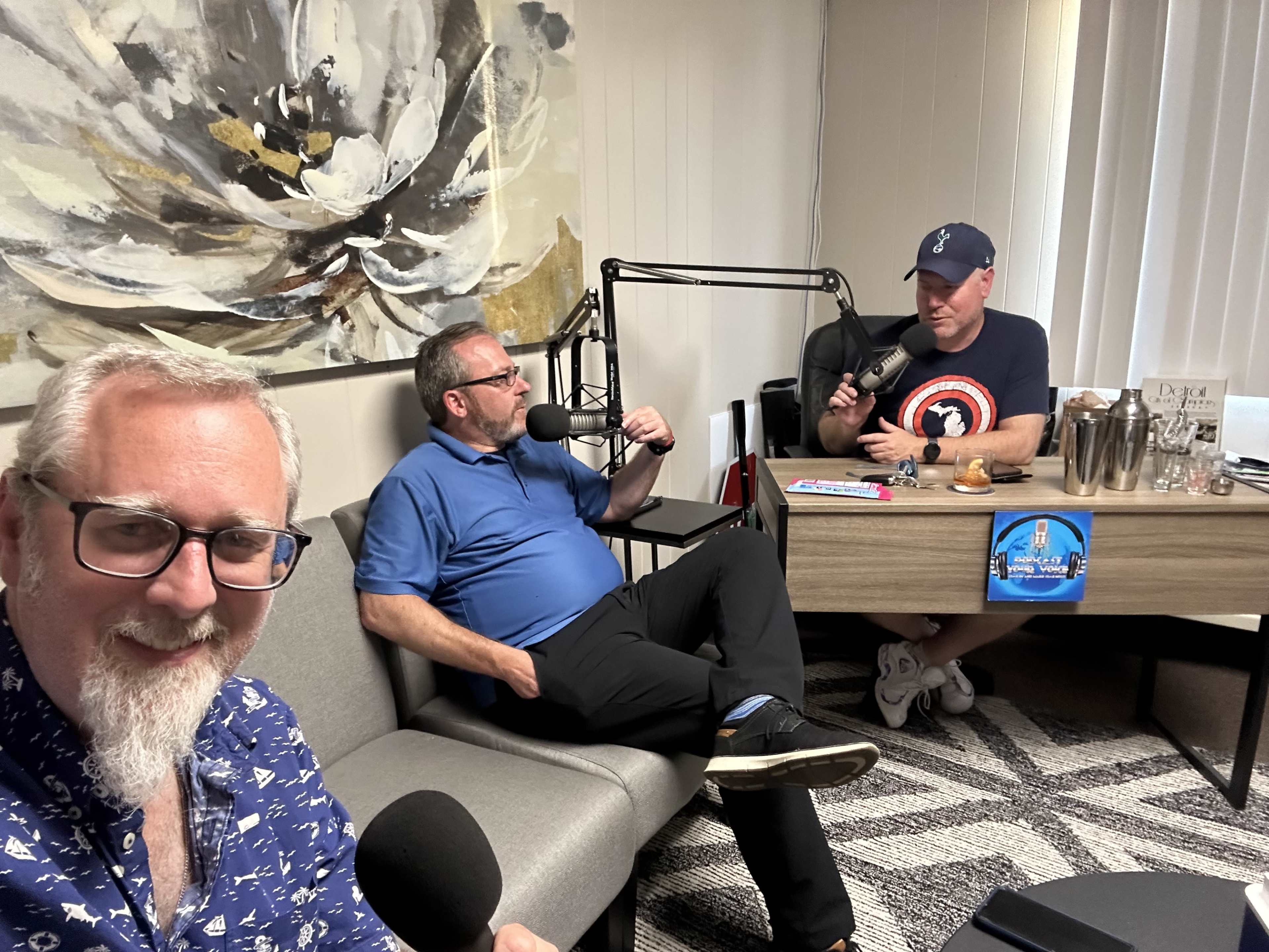 Three men are engaged in a podcast discussion in a modern office setting, with one man holding a microphone while seated at a desk, and the other two are on a gray couch, one posing for a selfie.