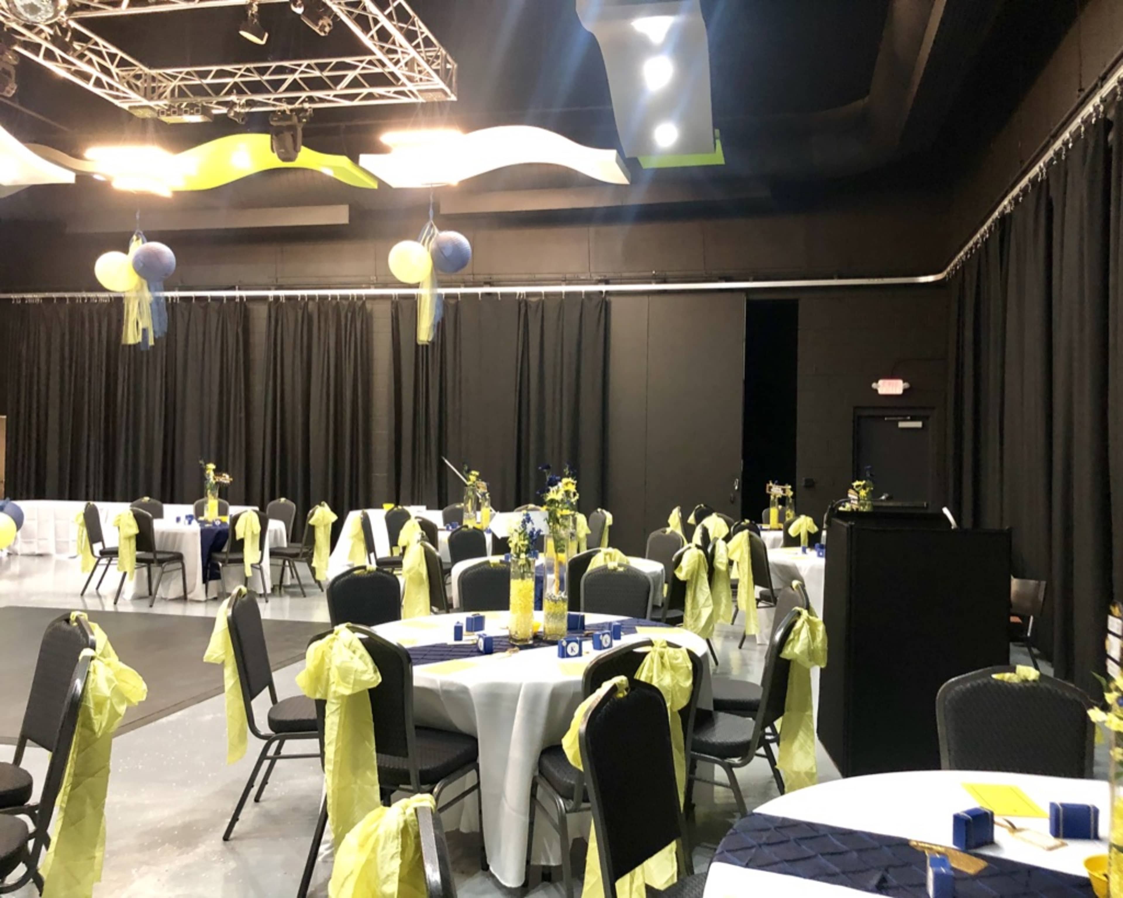 A banquet hall set up for an event, featuring round tables adorned with yellow tablecloths and blue decorations, surrounded by black chairs with yellow covers.