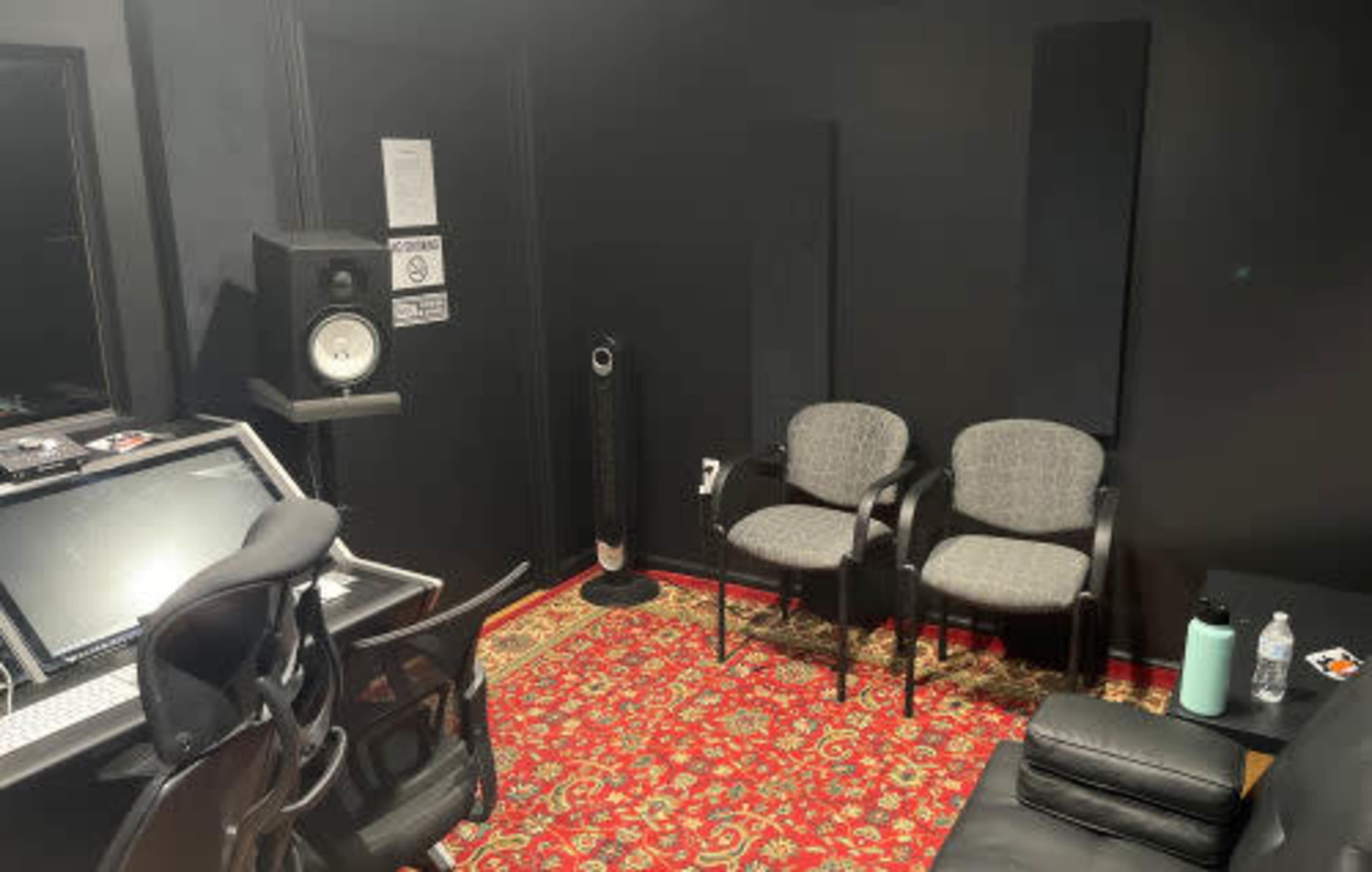 A small, dimly lit recording studio with a carpeted floor, two chairs, a desk with audio equipment, and soundproofing panels on the walls.