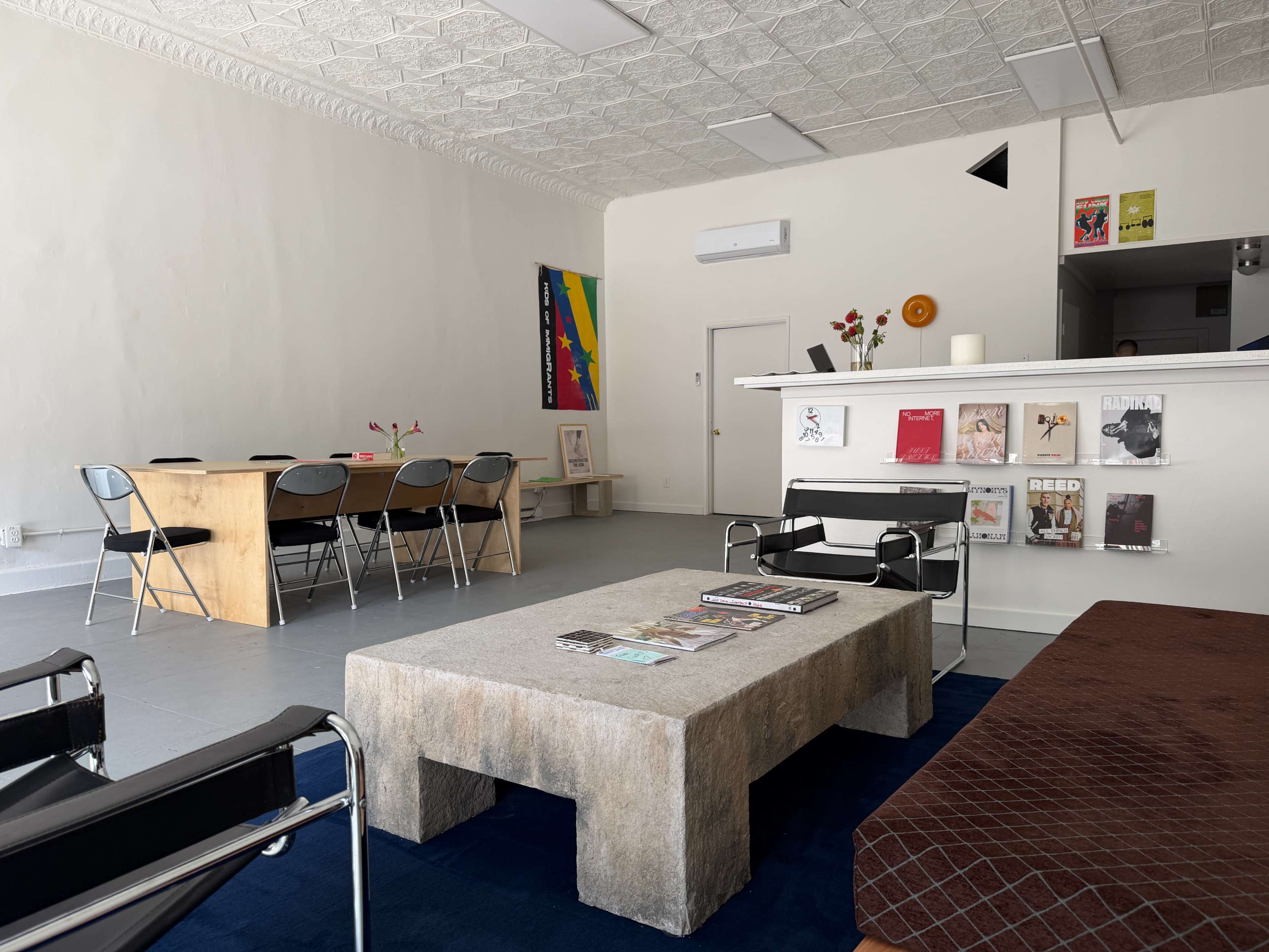 The image shows a minimalist interior of a room featuring a rectangular coffee table, a seating area with chairs, and a wall displaying magazines and posters.