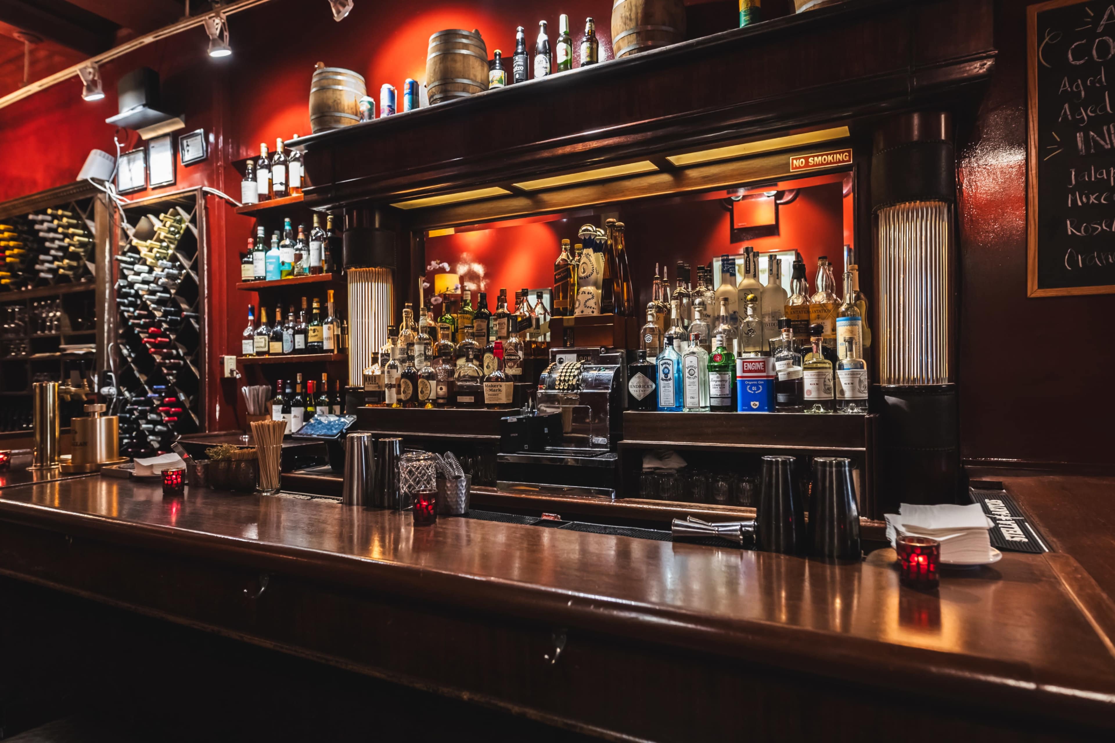 The image shows a wooden bar with various bottles of liquor displayed on shelves and behind the bar, set against a red wall adorned with decorative elements.