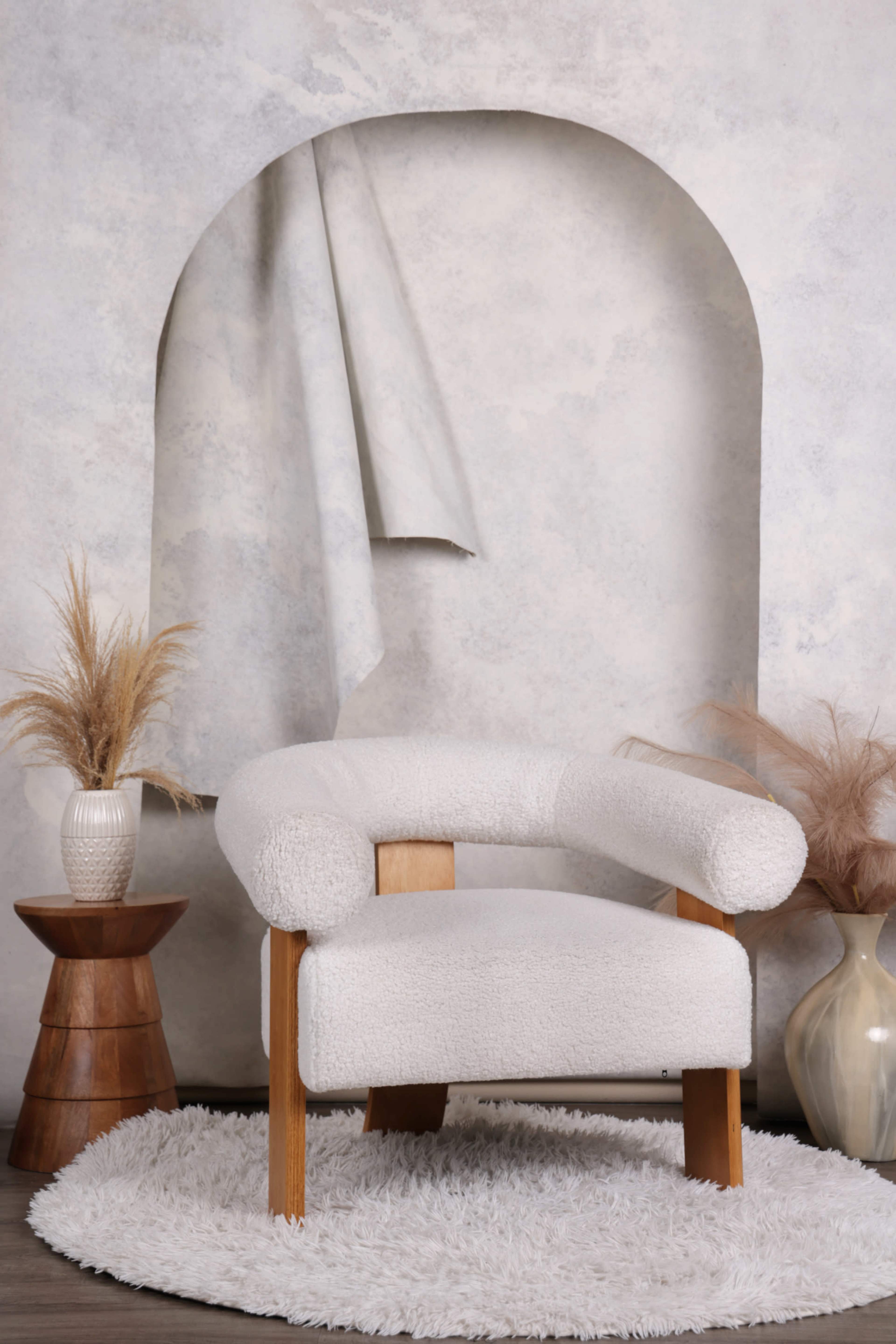 A plush white chair with wooden legs is positioned in front of a textured wall, accompanied by two decorative vases and a textured rug.