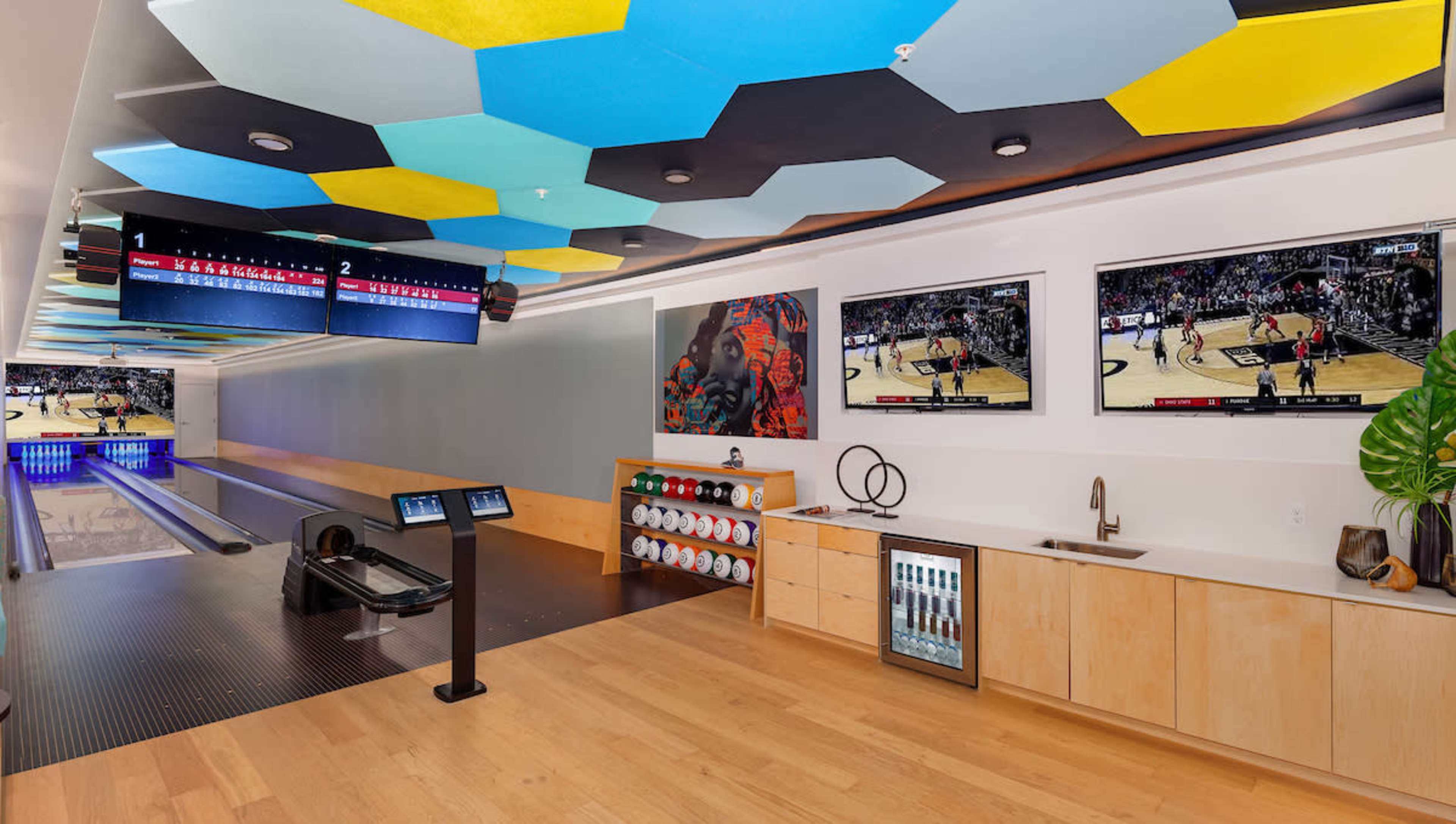 A modern bowling alley with two lanes, a seating area, two large televisions displaying a basketball game, and a colorful hexagonal ceiling.