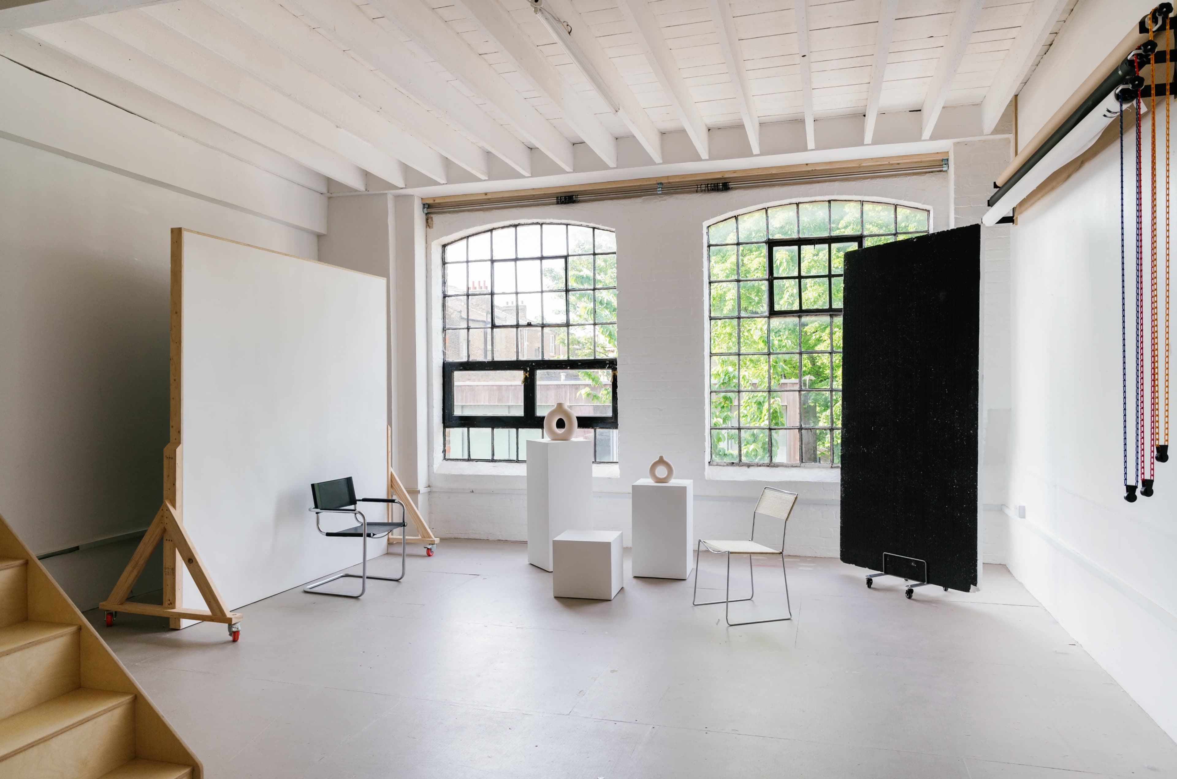 A spacious, well-lit studio featuring three white pedestals, two chairs, and large windows that overlook greenery.