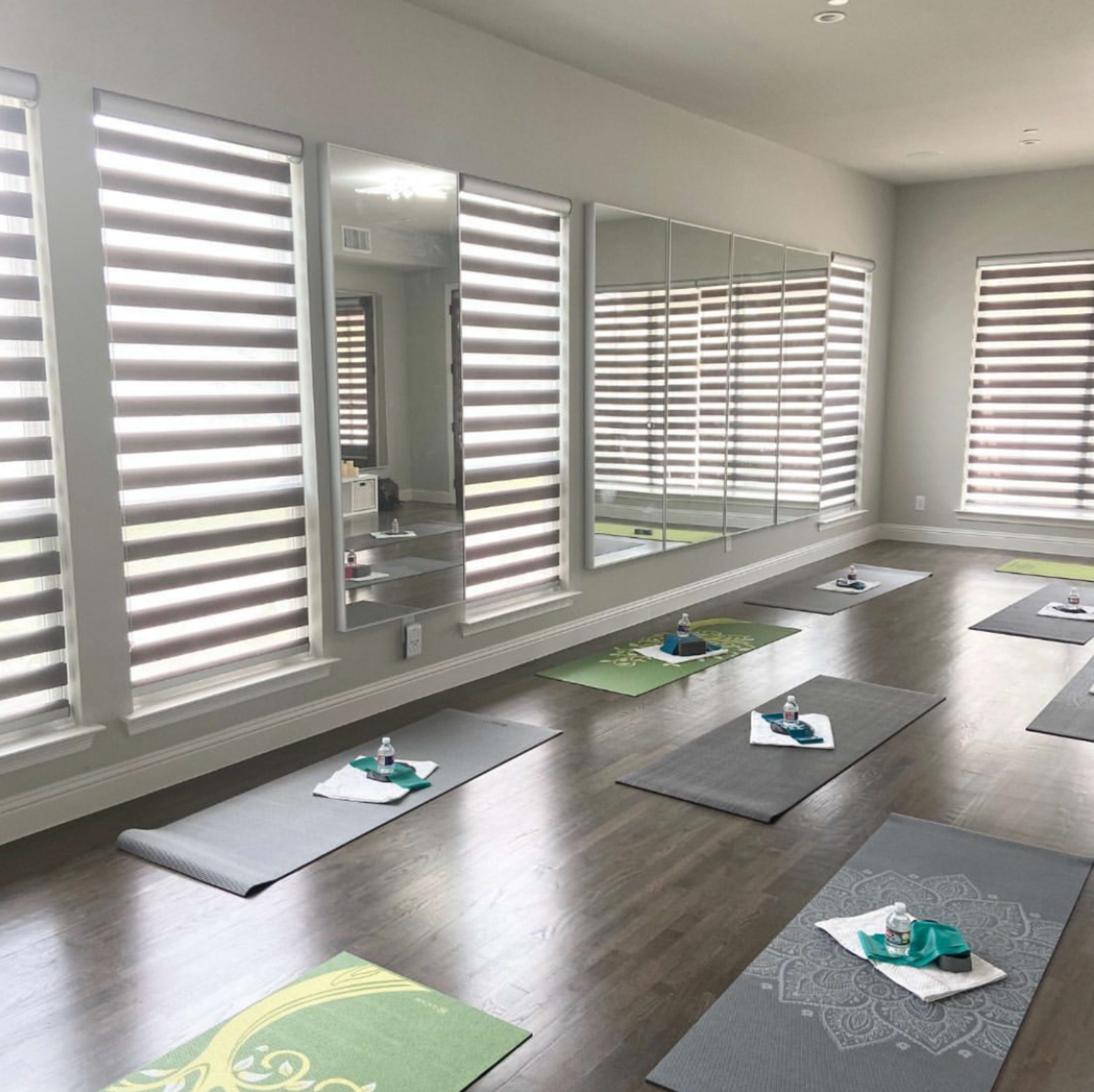 The image shows a yoga studio with several exercise mats arranged on a wooden floor, reflecting daylight through striped window shades.