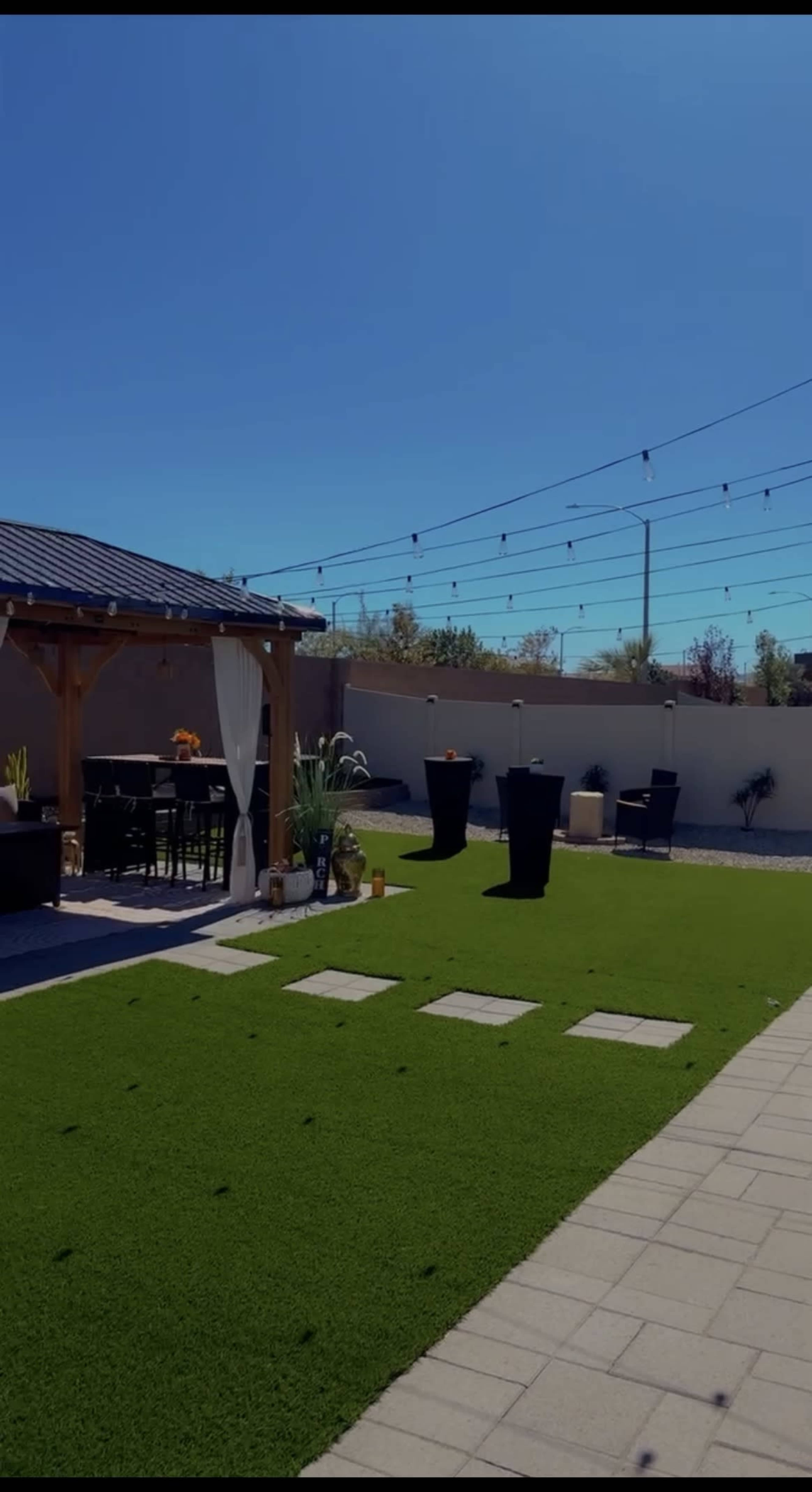 The scene features a landscaped backyard with artificial grass, a wooden pergola, outdoor seating, and garden lights strung overhead.
