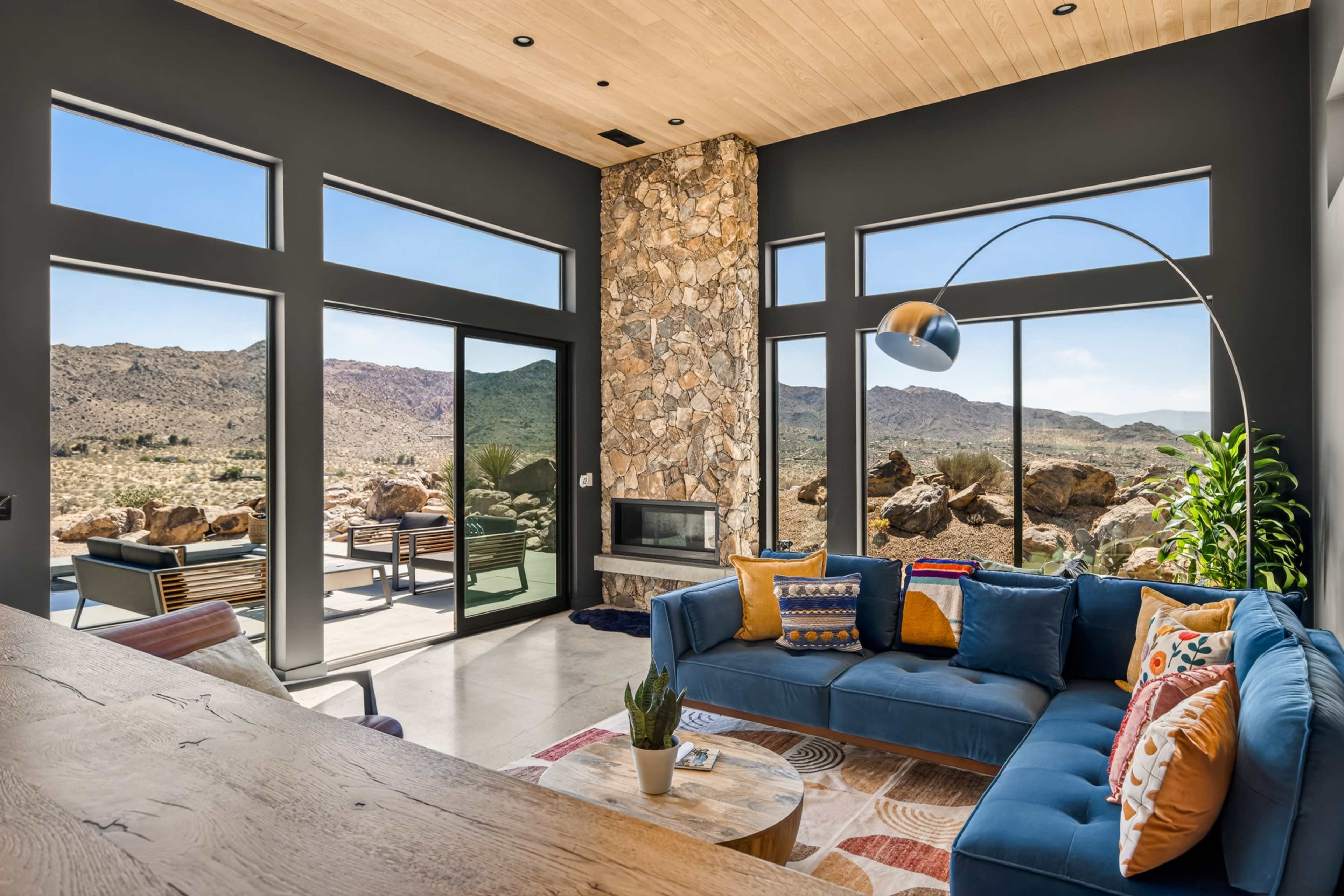 The image shows a modern living room with large windows overlooking a desert landscape, featuring a blue sectional sofa, a stone wall, and minimalistic decor.