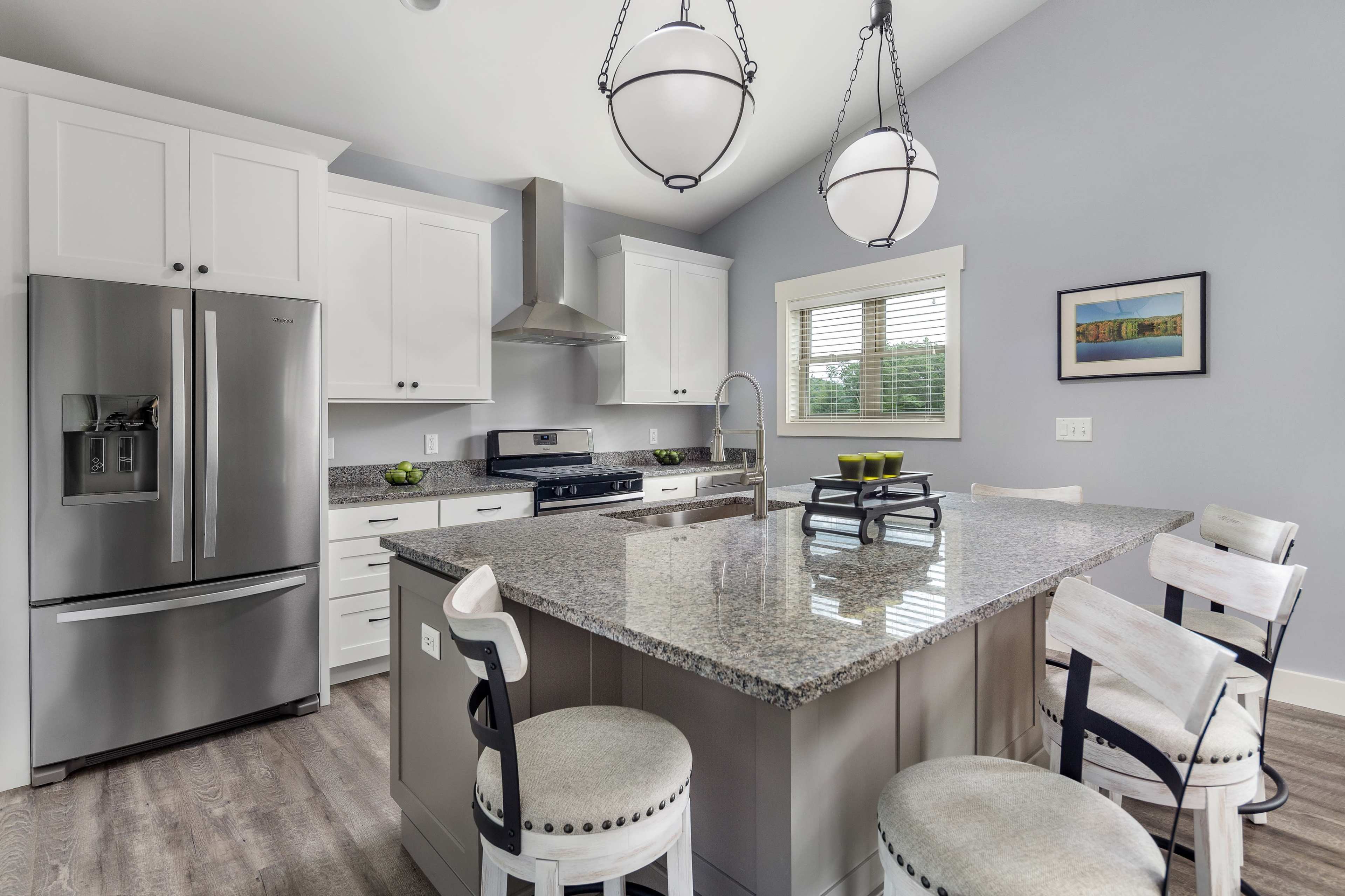 A modern kitchen features gray cabinets, a large granite island with seating, and stainless steel appliances.