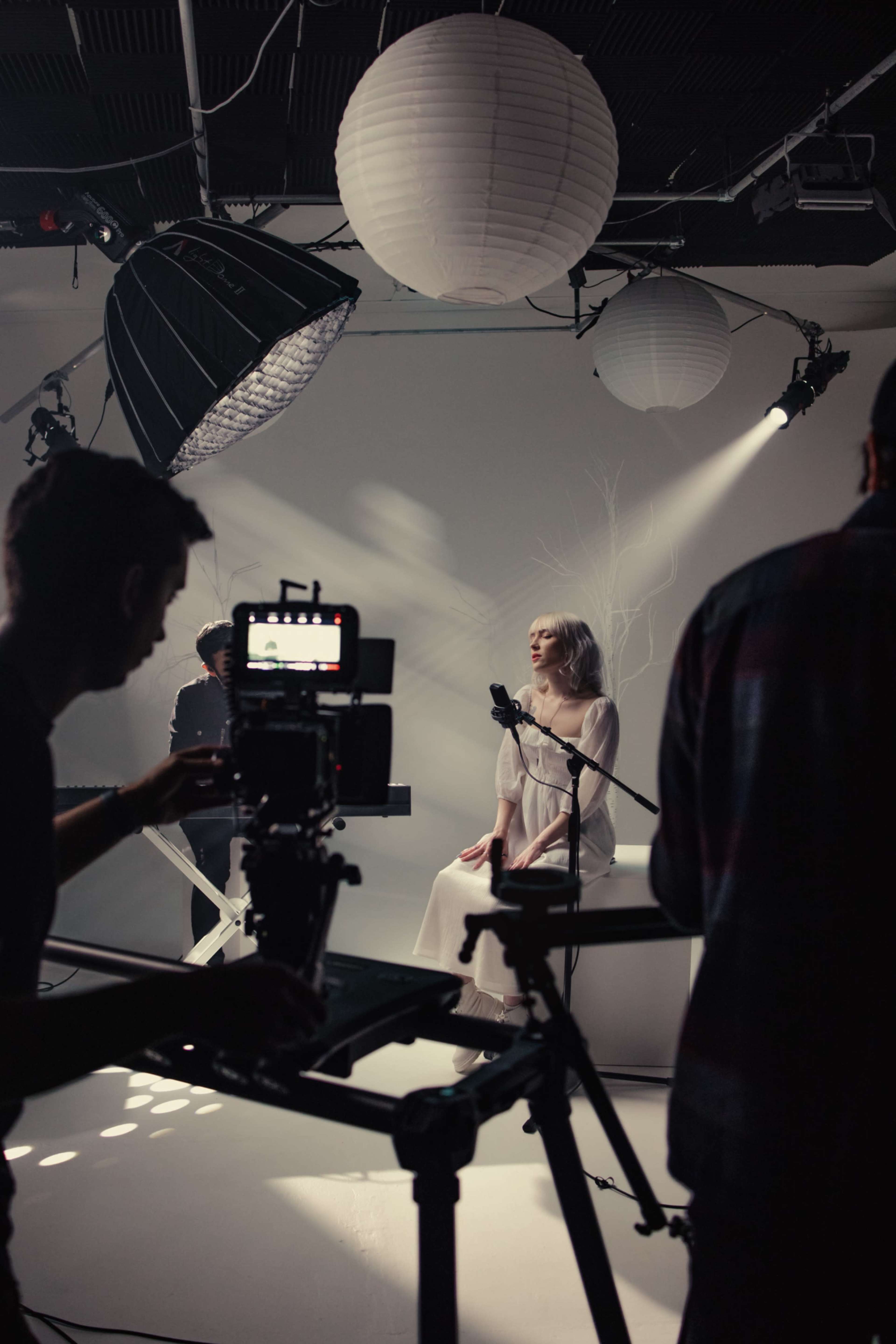 A female singer sits at a microphone on a small stage while a camera and lighting setup are positioned around her in a studio.