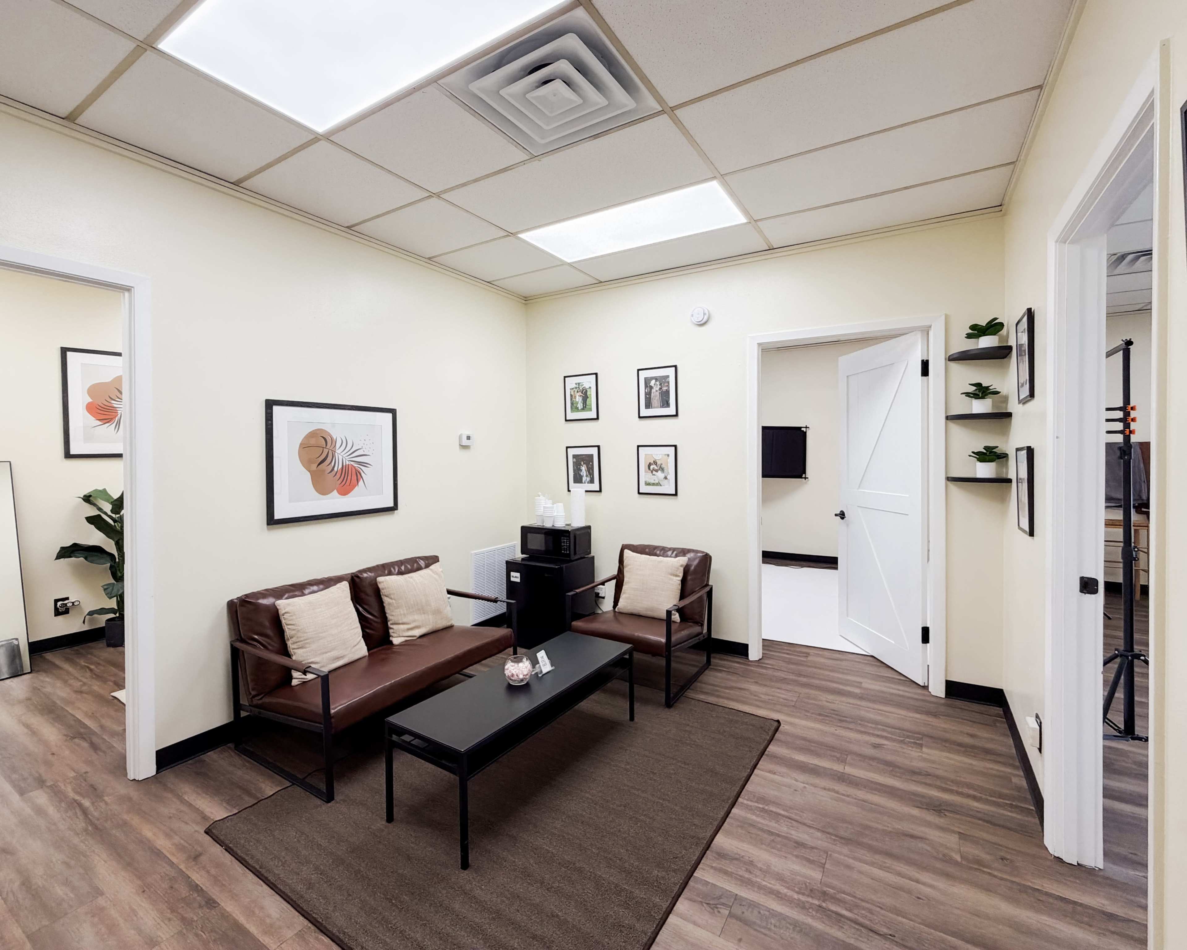 The image shows a small waiting room with two brown leather sofas, a coffee table, framed artwork on the walls, and a plant in the corner.