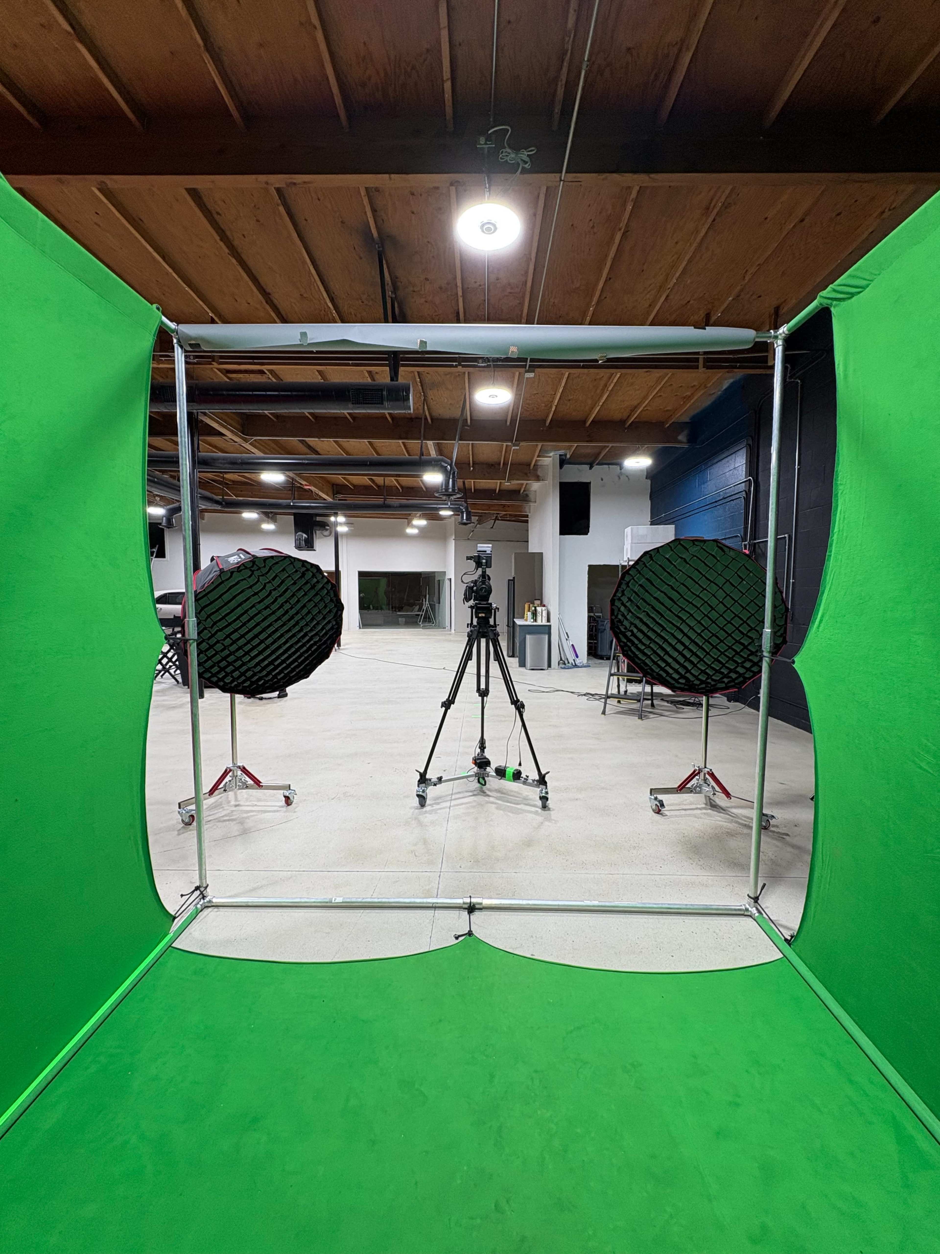 The image shows a green screen setup in a spacious studio with a camera on a tripod positioned in the center and two light softboxes on either side.
