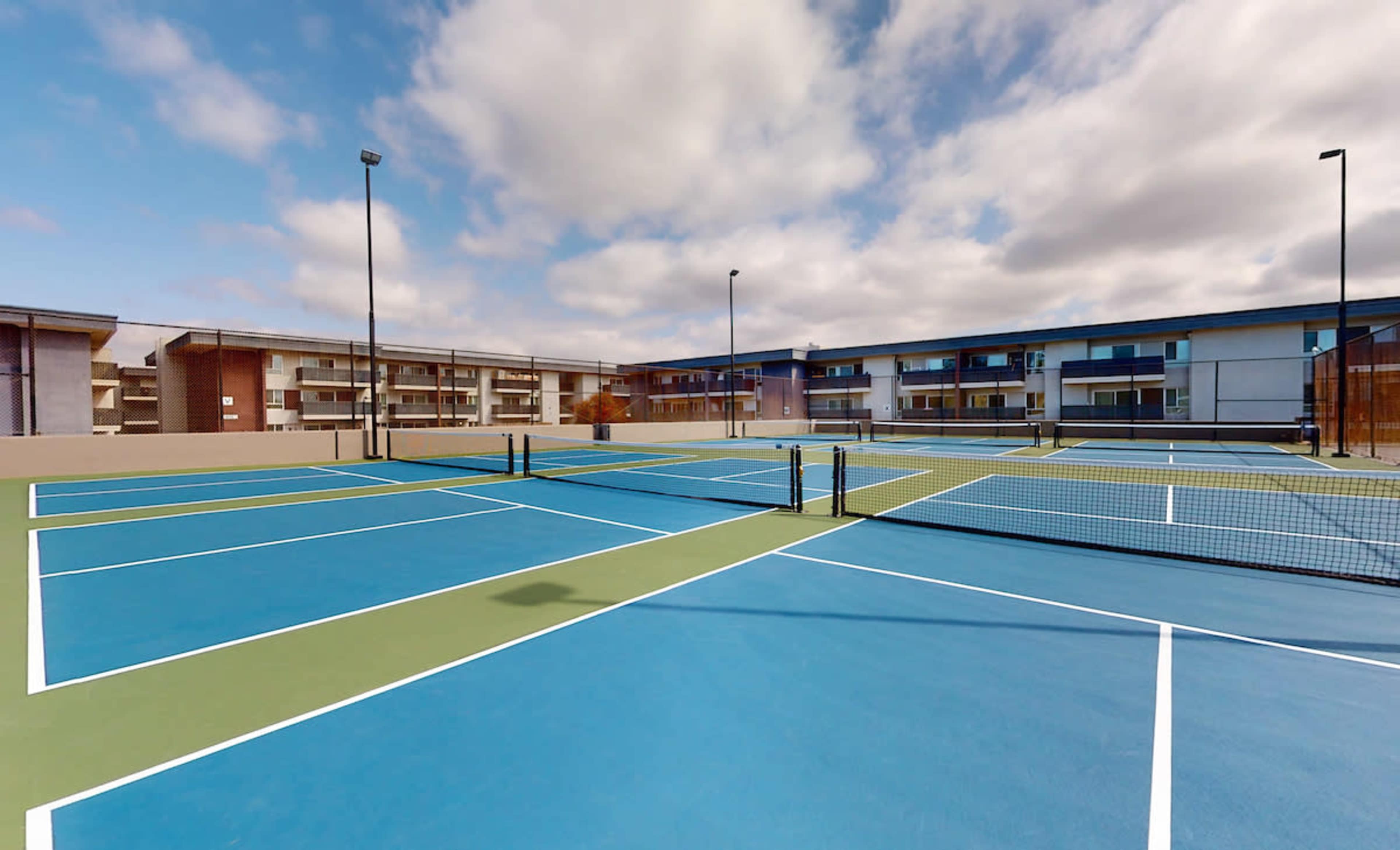 A series of blue and green tennis courts situated near a multi-story building under a partly cloudy sky.