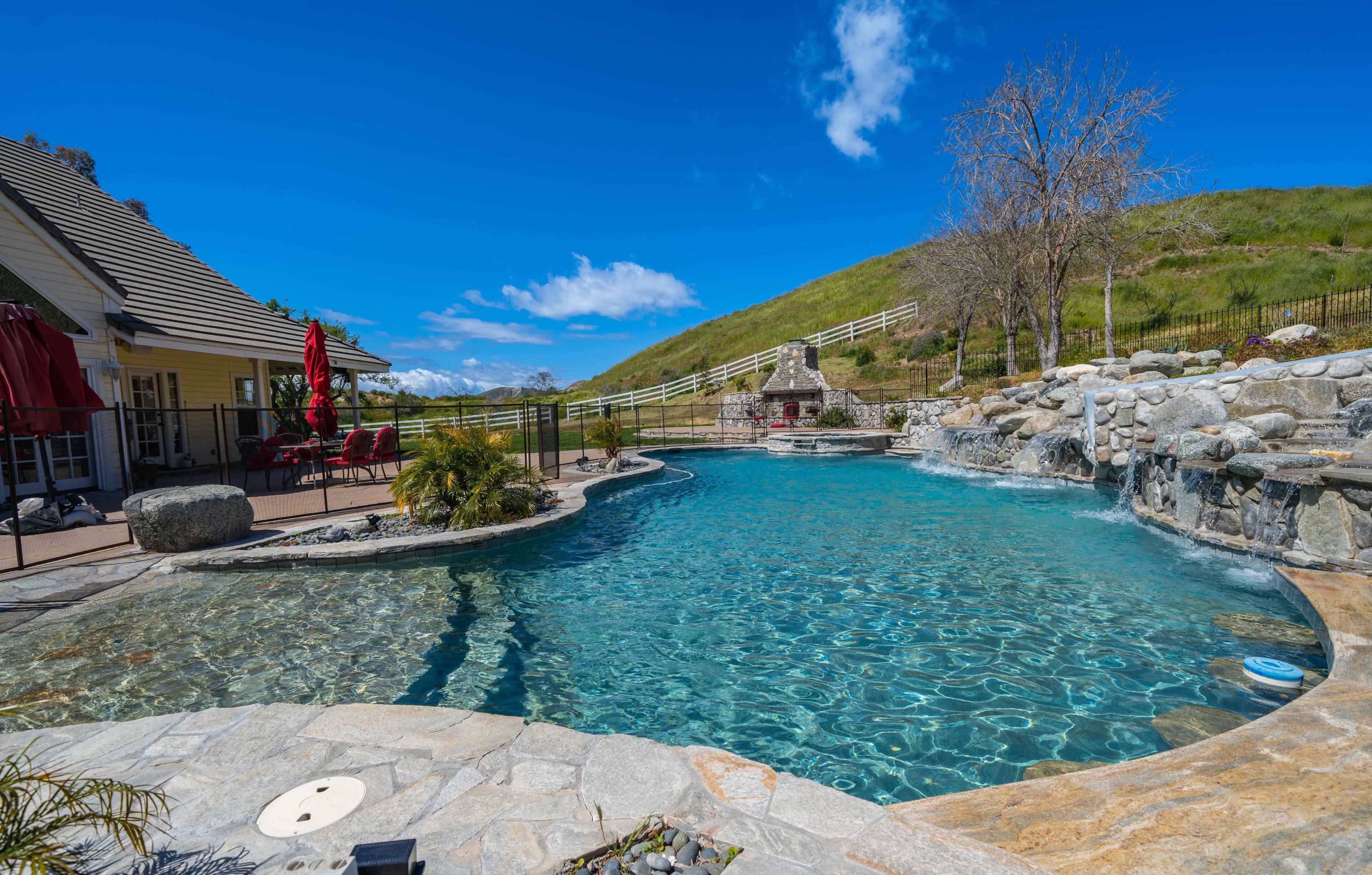 The image shows a curved swimming pool surrounded by stone patio, a small waterfall feature, and a green hillside in the background.